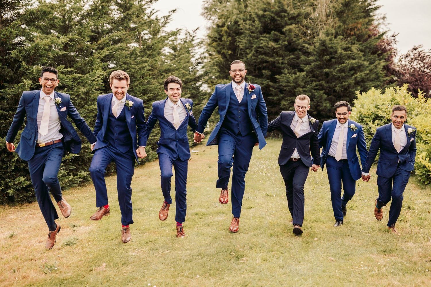 A group in matching navy suits leaps joyfully together on a grassy lawn with evergreen trees in the background.