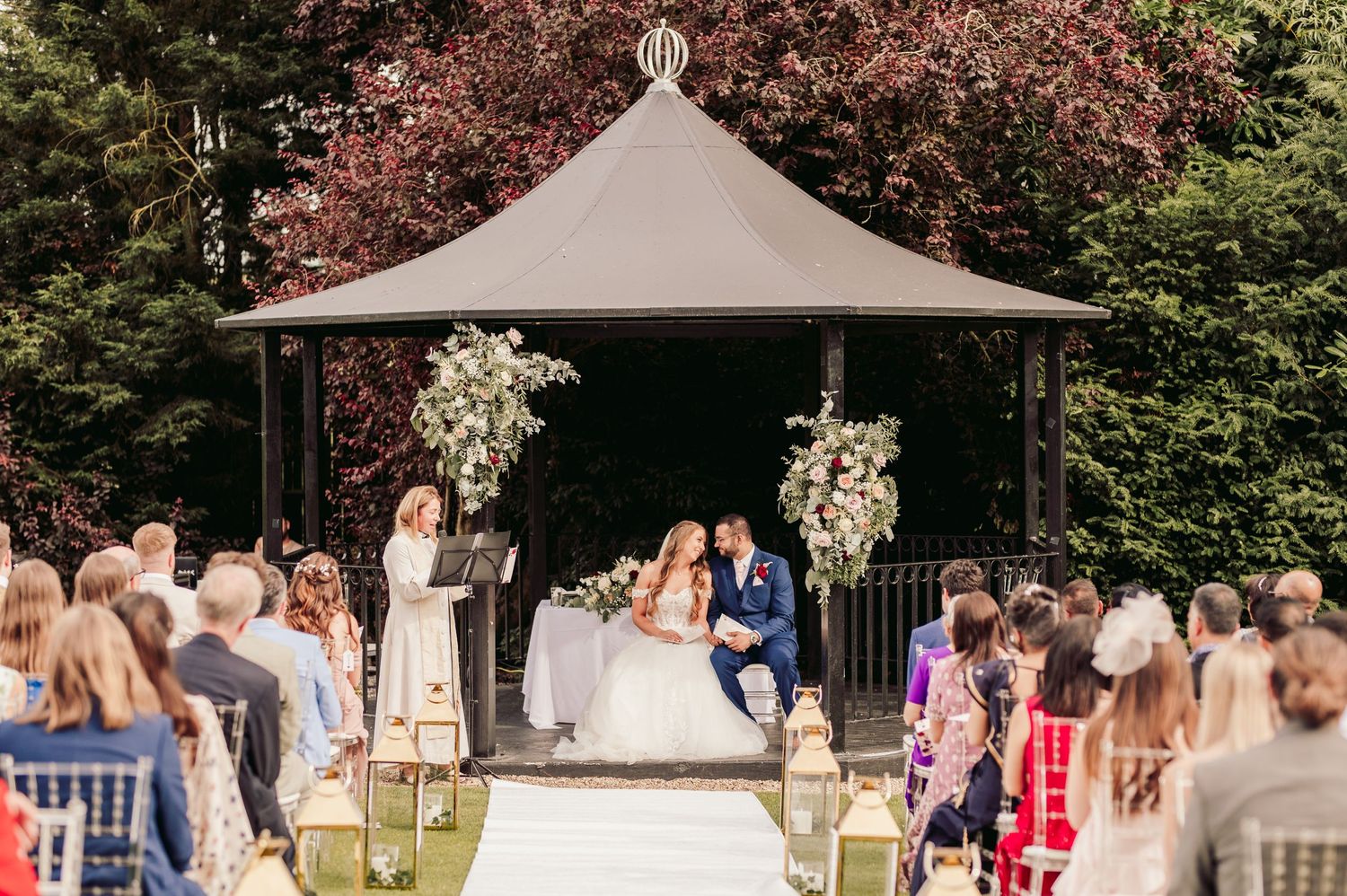 An outdoor wedding ceremony takes place under an elegant gazebo decorated with white floral arrangements.