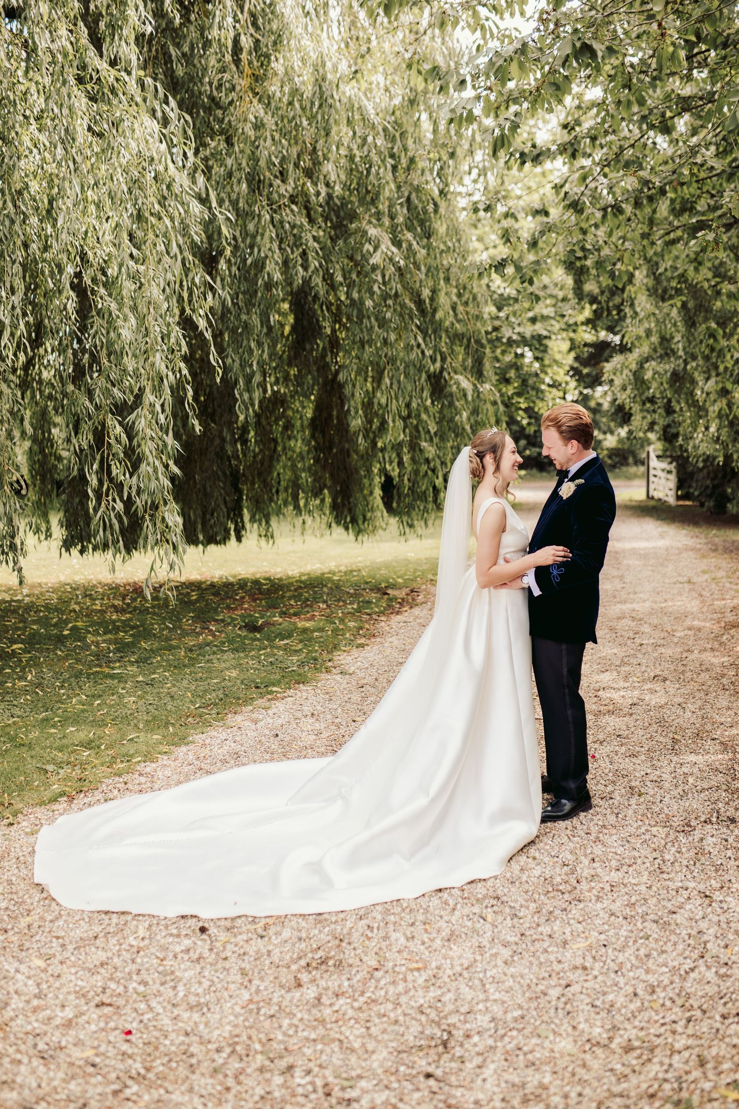 A couple shares an intimate moment under a weeping willow tree on a gravel path in an elegant wedding setting.