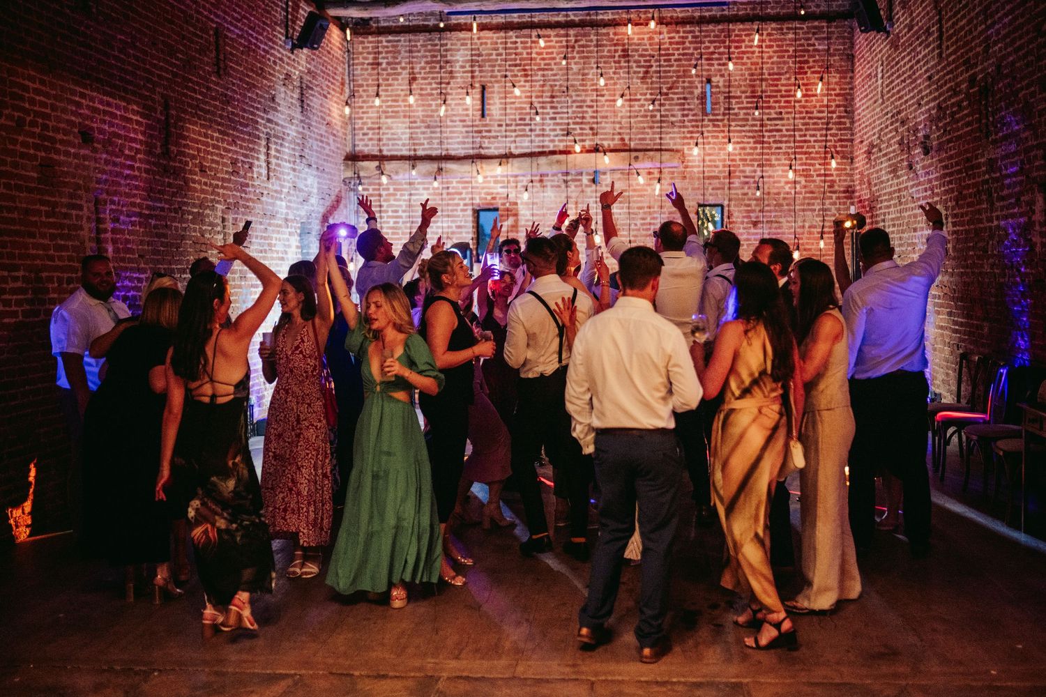 Wedding guests dance and celebrate on a rustic brick-walled dance floor at an evening reception party.