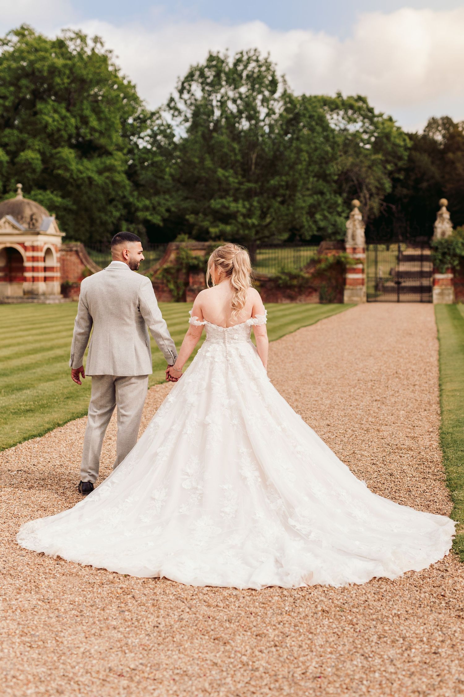 Elegant wedding couple walking on a gravel path with the bride's lace dress train trailing behind.
