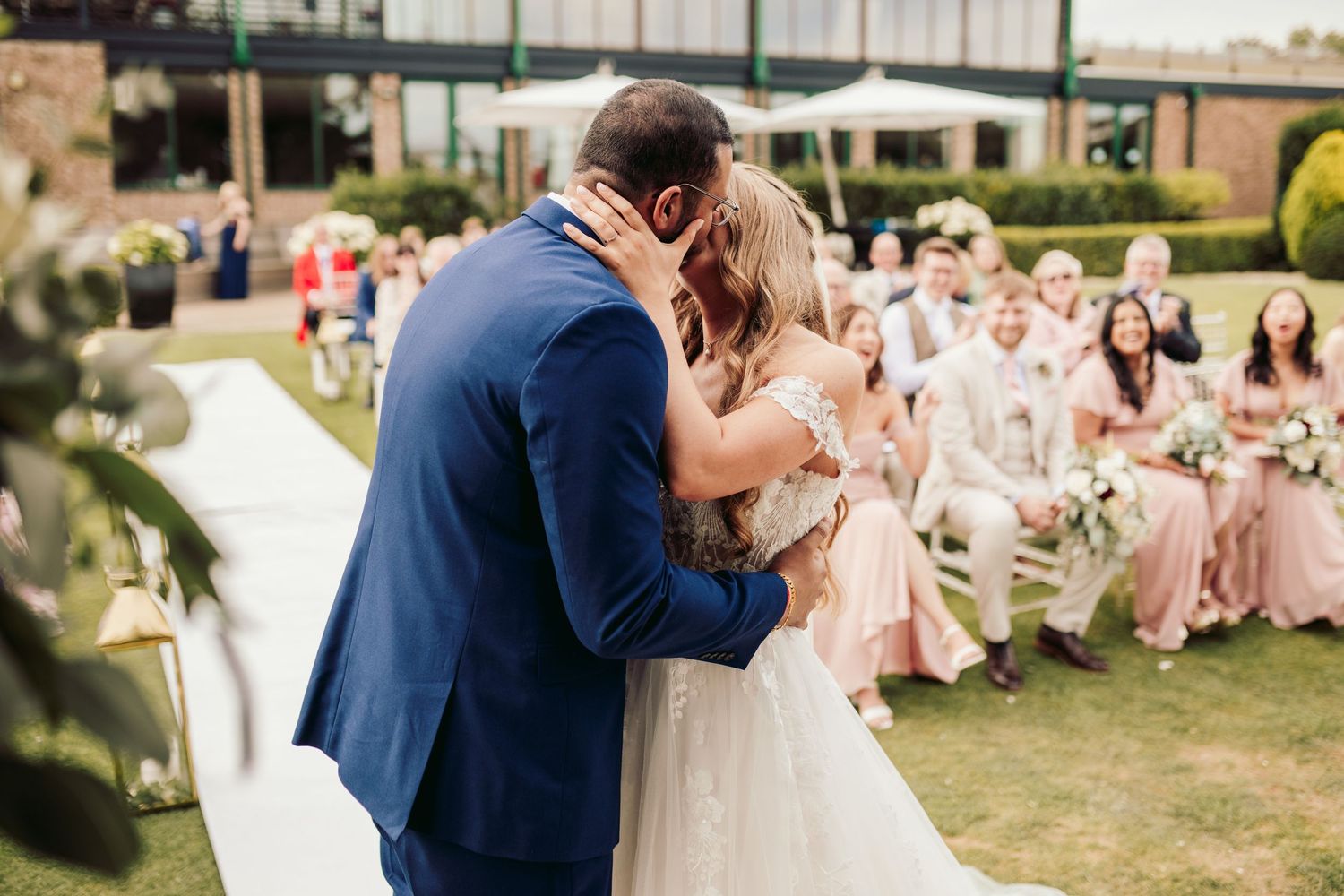 A couple shares a kiss at an outdoor wedding ceremony while guests in pink dresses look on from their seats on the lawn.