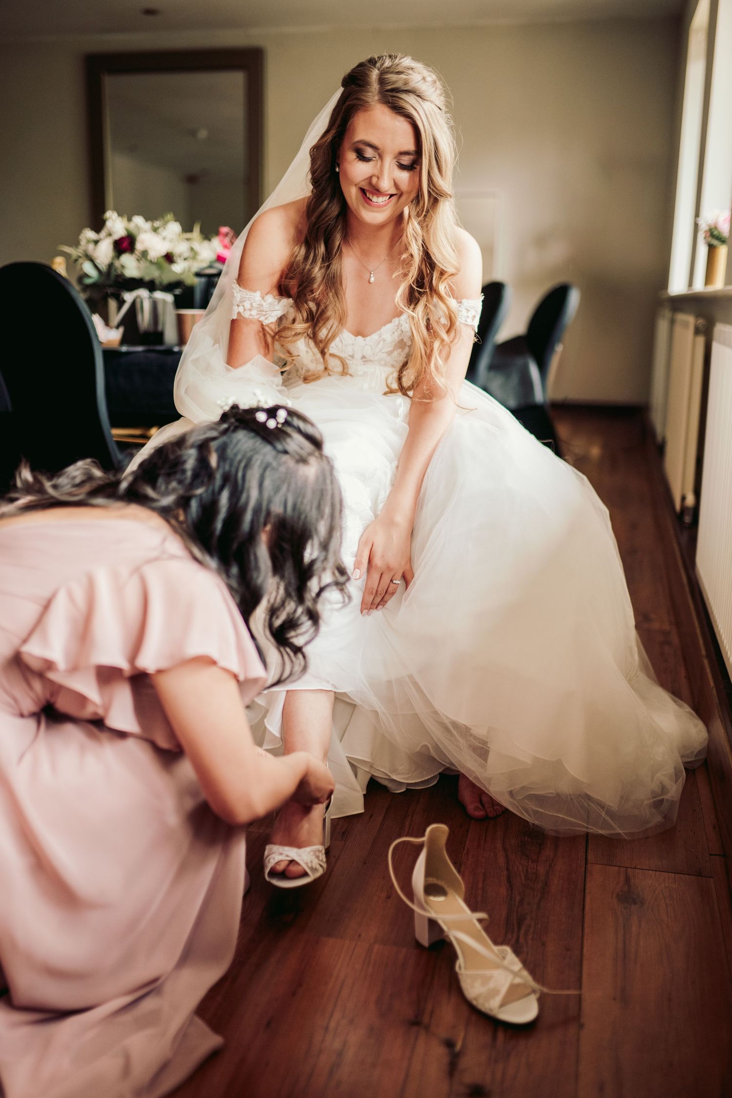A bride in a white wedding dress smiles while her bridesmaid in pink helps with wedding shoes on hardwood floor.