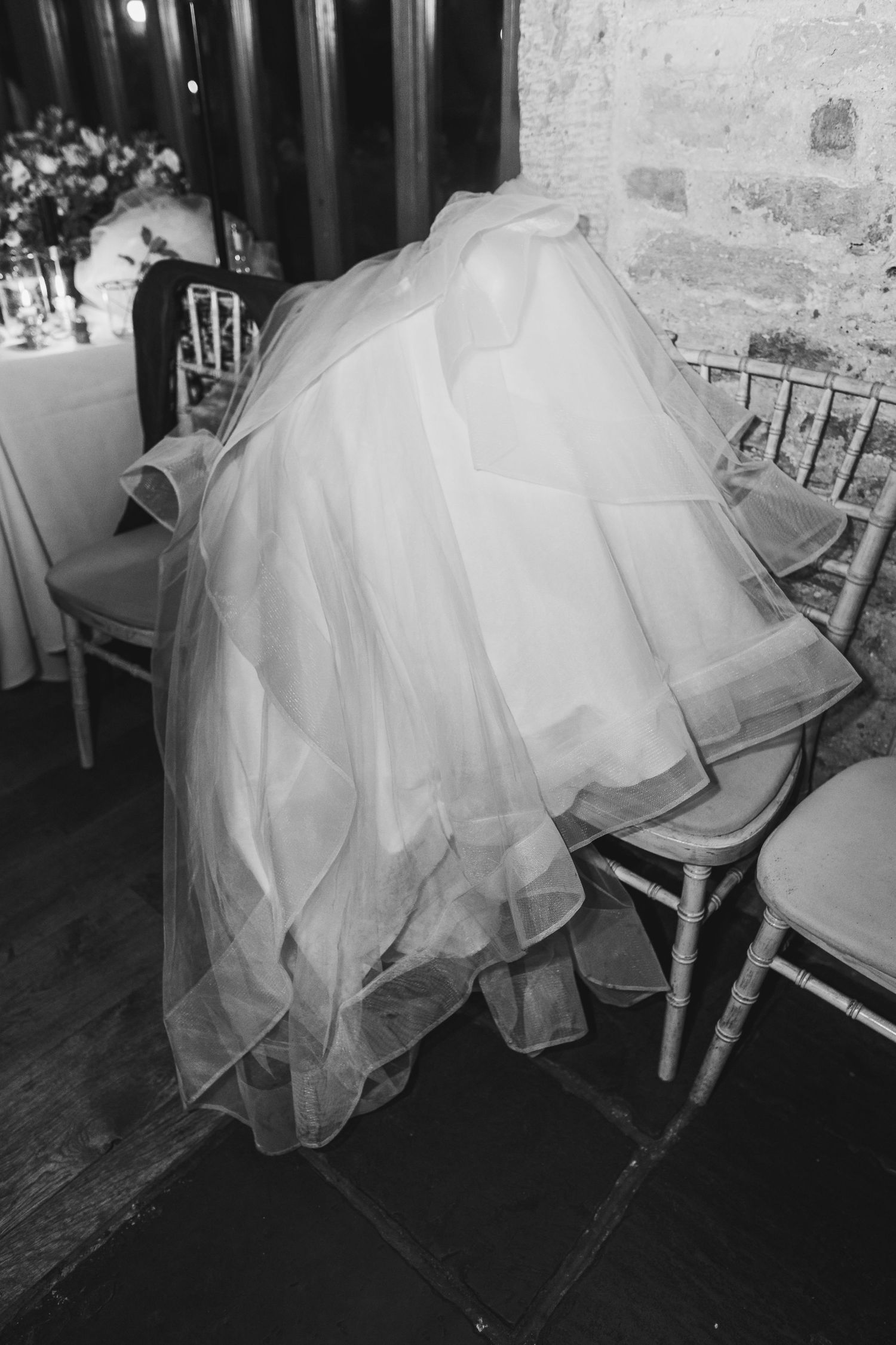 Black and white photo of a flowing wedding dress skirt against wooden chairs.