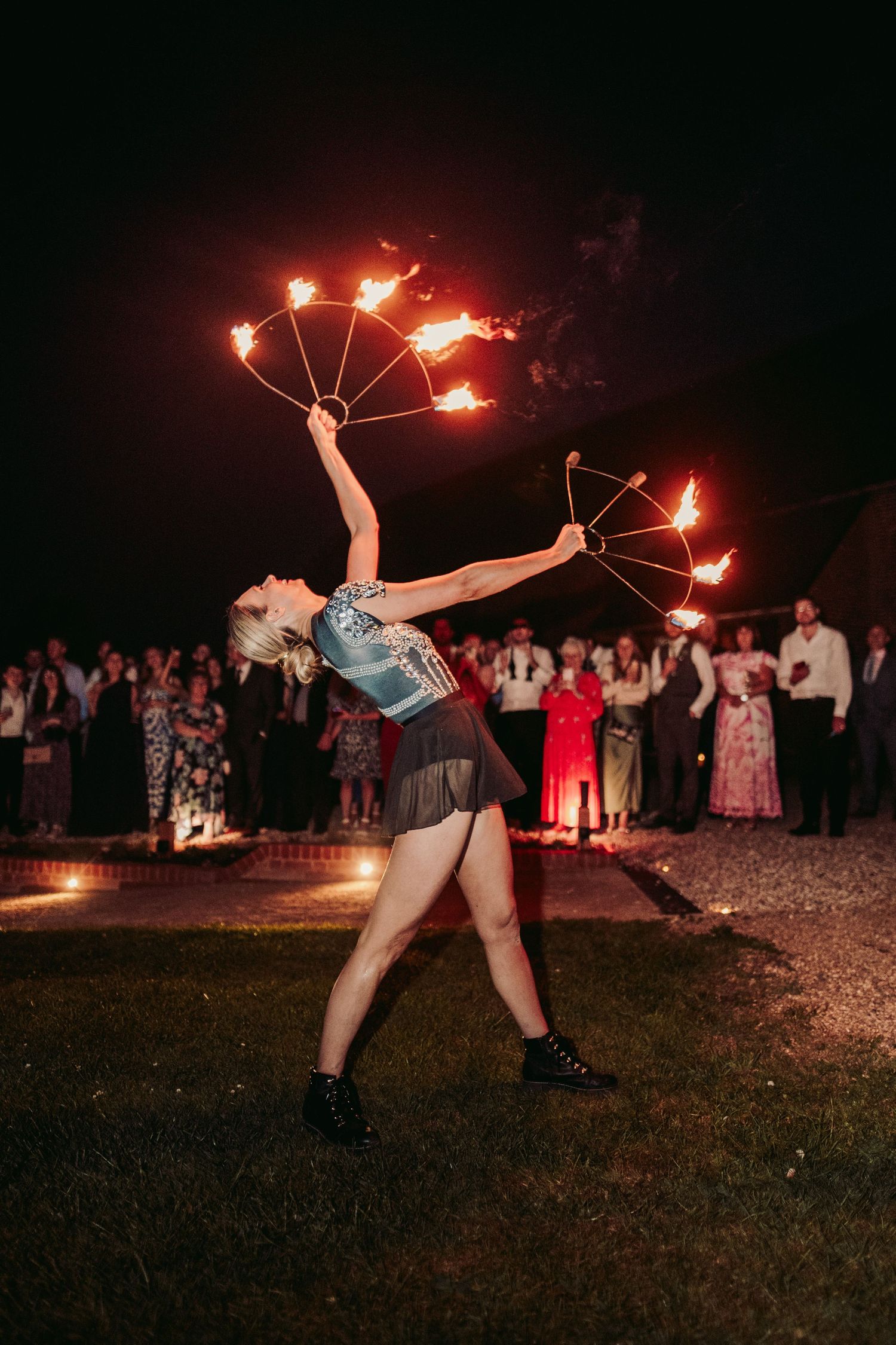 Fire performer in denim shorts and boots spins flaming batons at night while audience watches in background.
