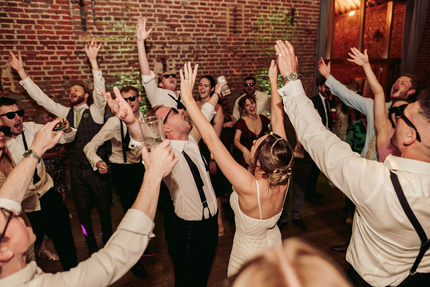 Wedding guests dance with raised arms on a crowded dance floor inside a rustic brick-walled venue.