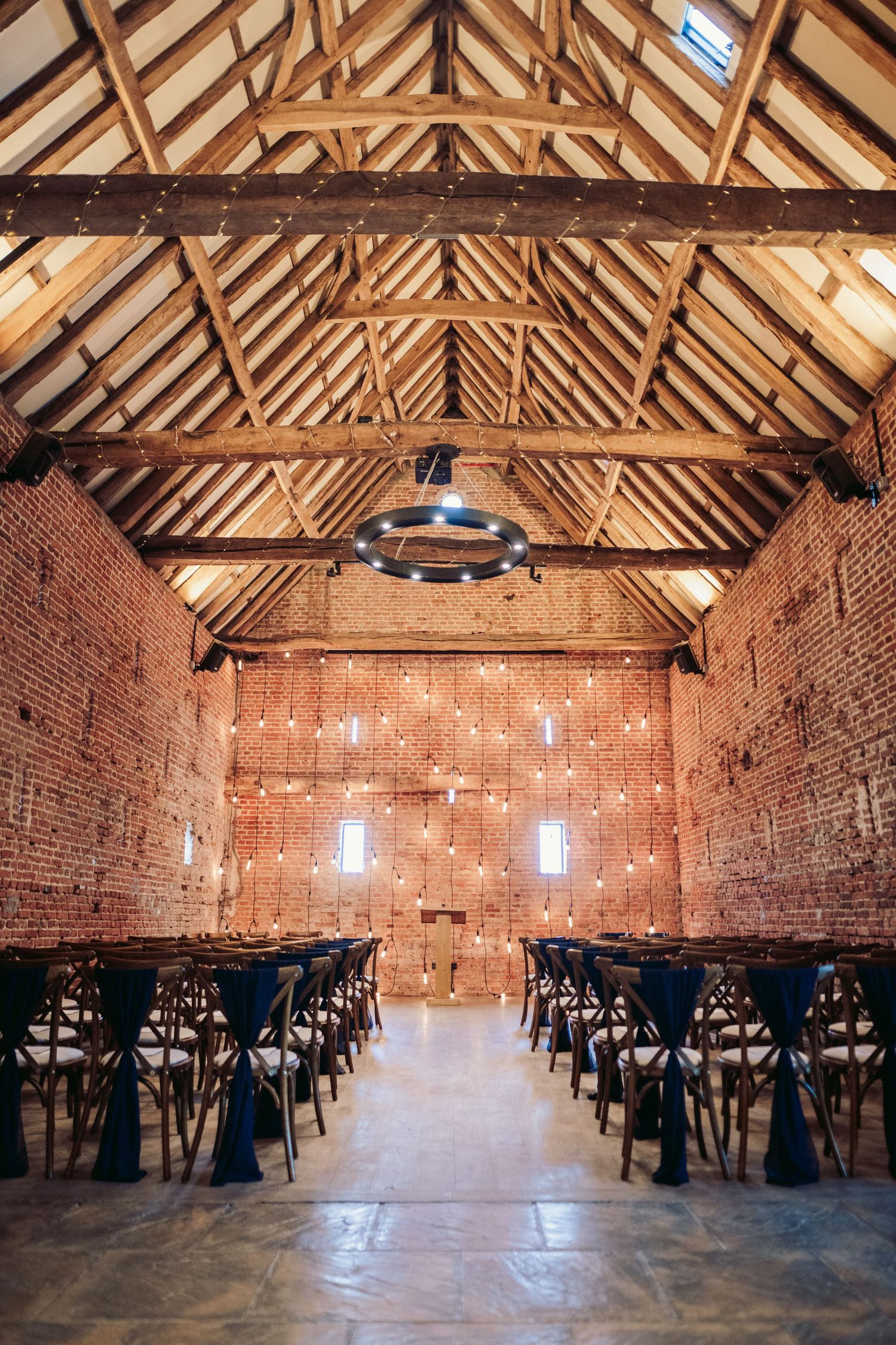 Rustic barn interior with exposed wooden beams and brick walls set up with rows of dark chairs for an event.