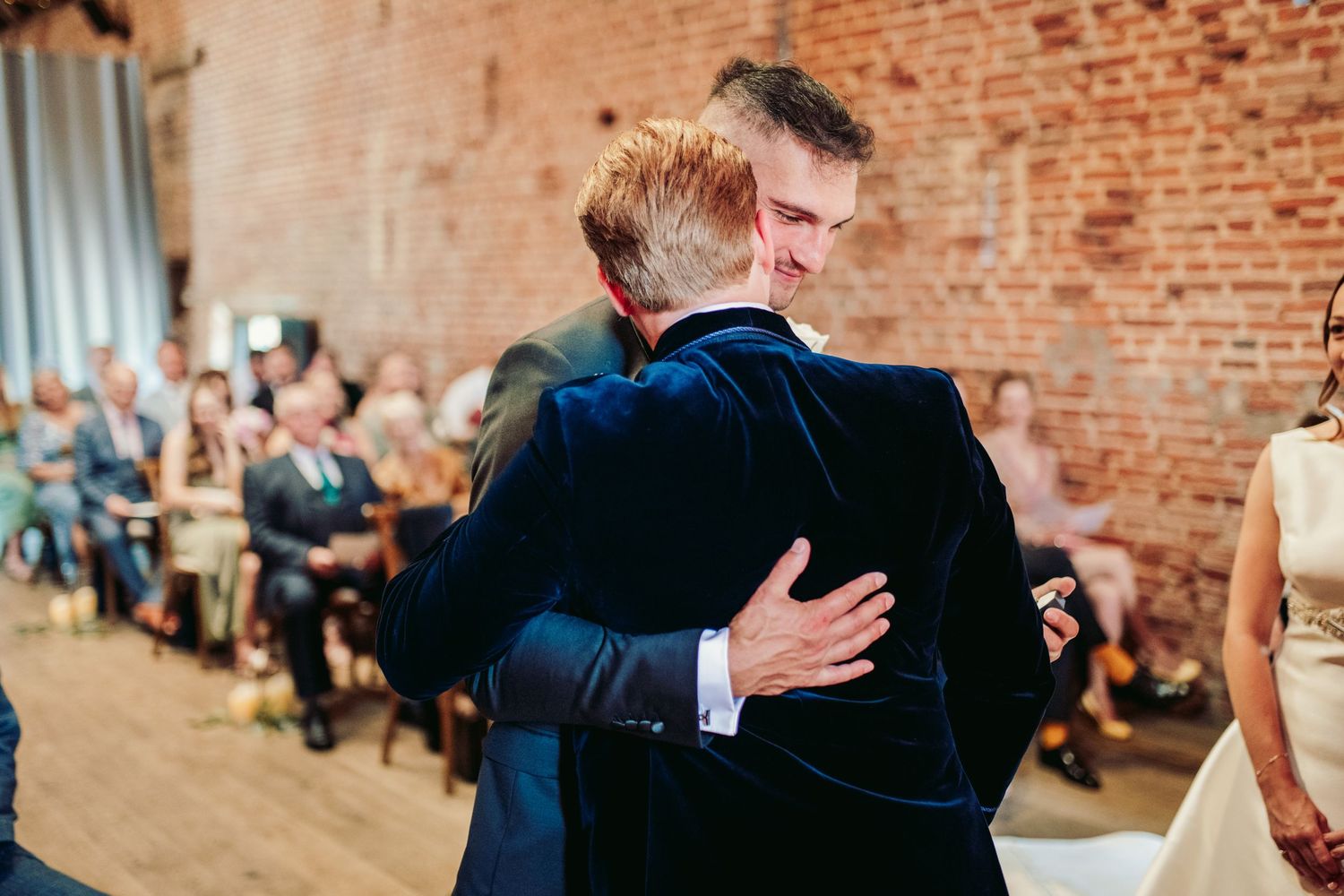 Two people share an emotional embrace during a wedding ceremony in a rustic brick-walled venue.