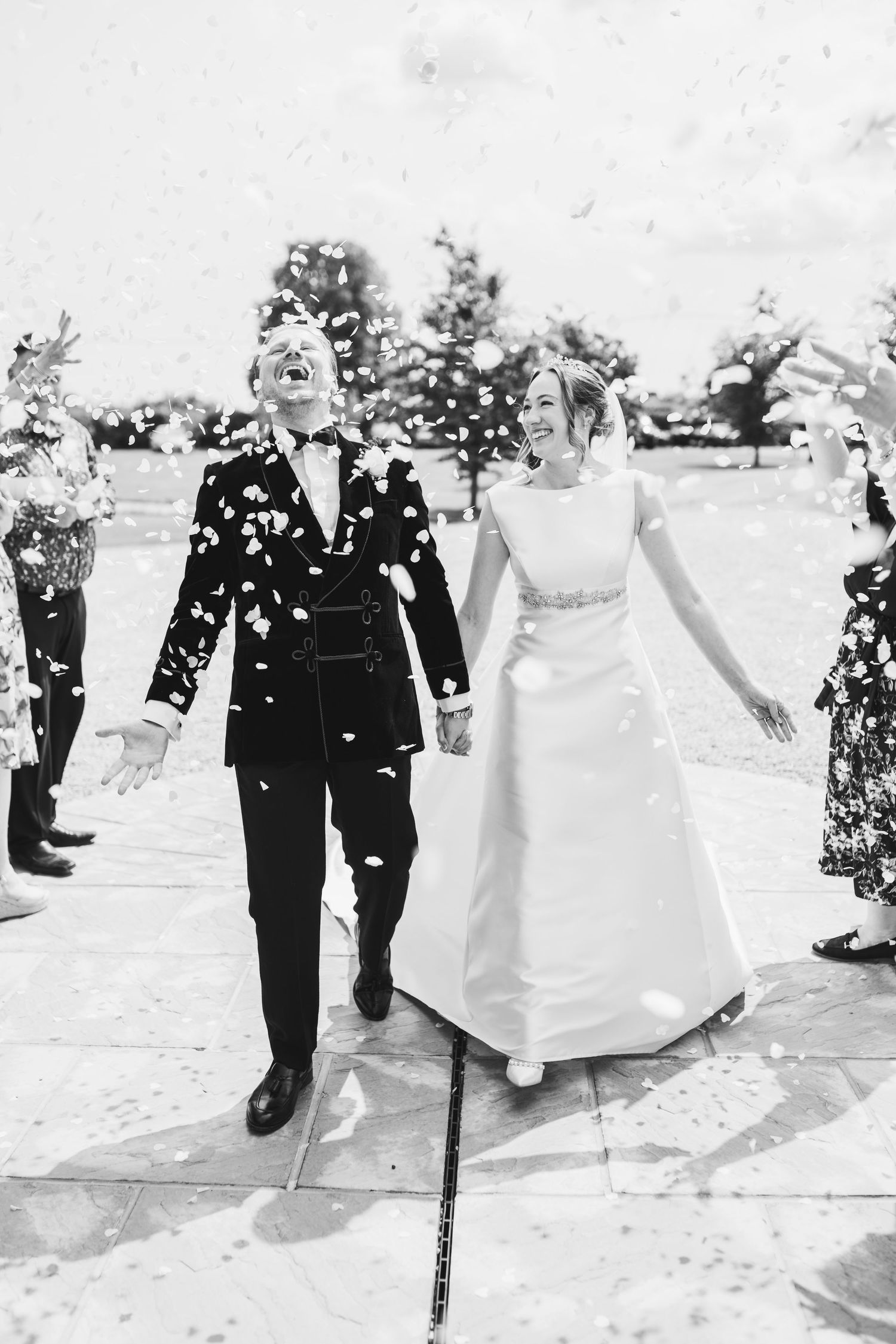 A black and white wedding photo showing a couple walking together while guests shower them with confetti outdoors.