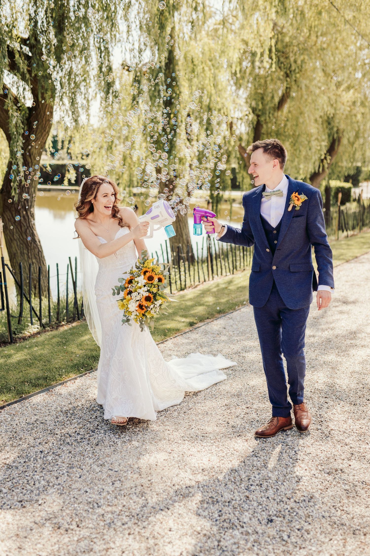 Newlyweds walk along a tree-lined gravel path beneath weeping willow trees.