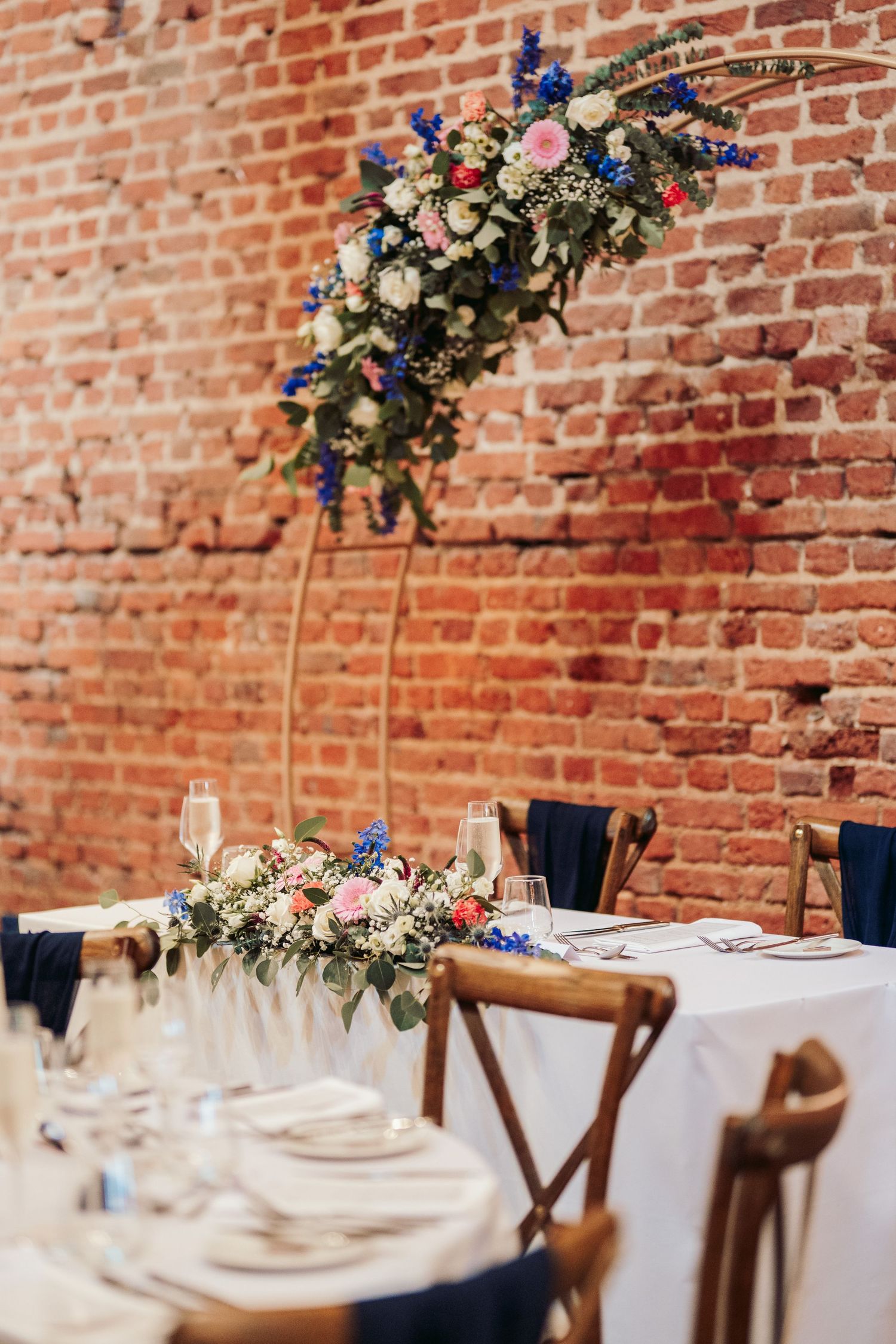 Elegant wedding table setting with floral arrangements against rustic brick wall featuring blue and white flowers.