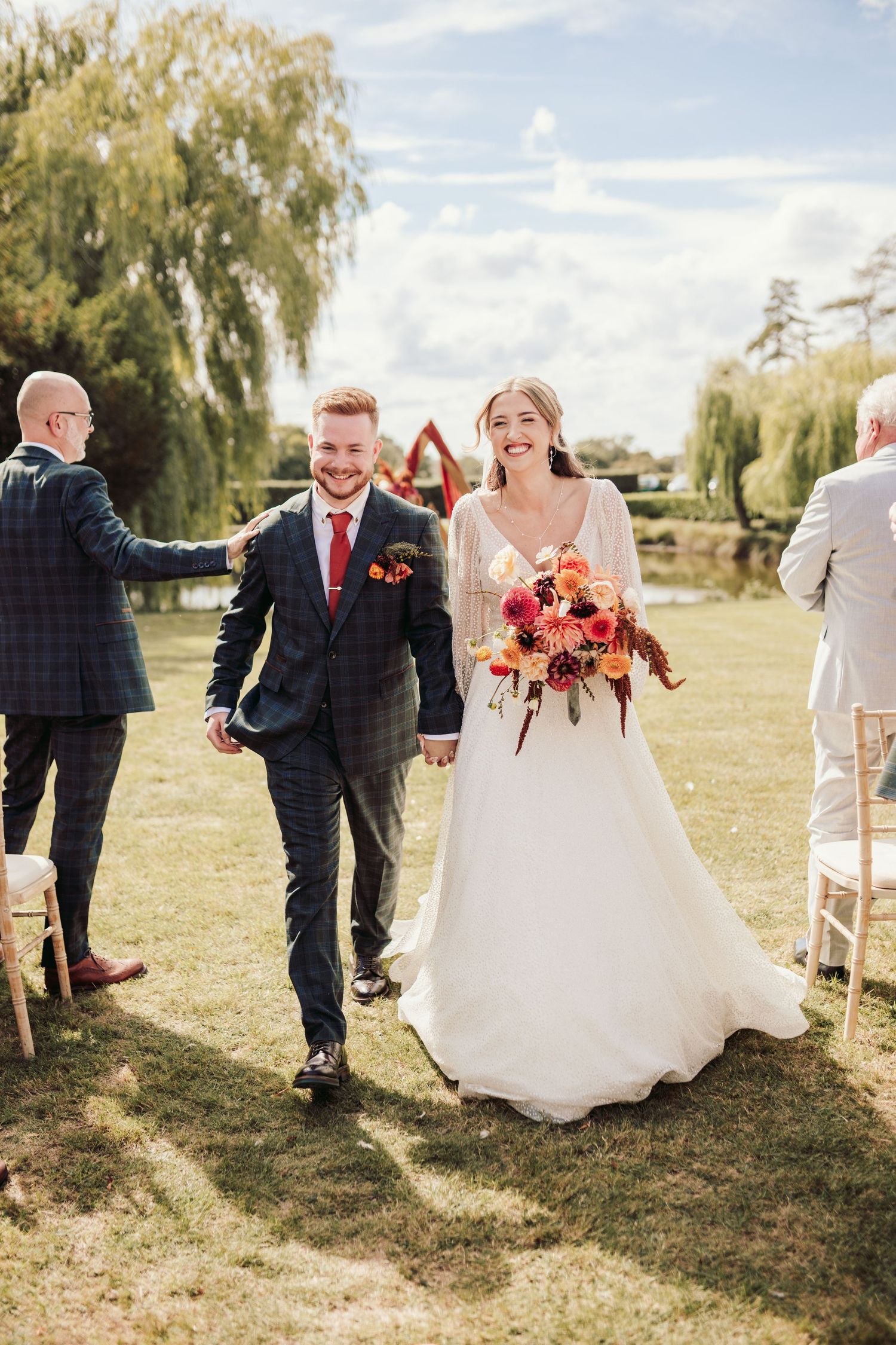 A joyful wedding couple walks outdoors on their special day with a stunning bouquet of red flowers.
