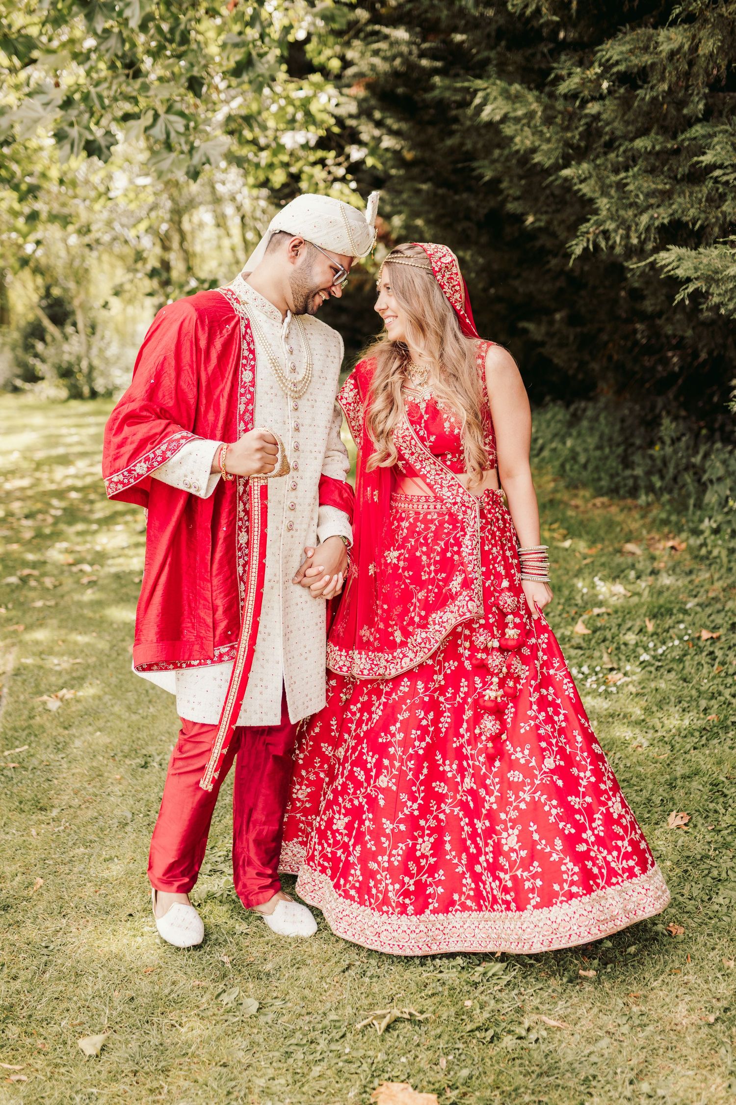 Traditional red and gold South Asian wedding attire shown in outdoor garden setting with natural sunlight filtering through trees.
