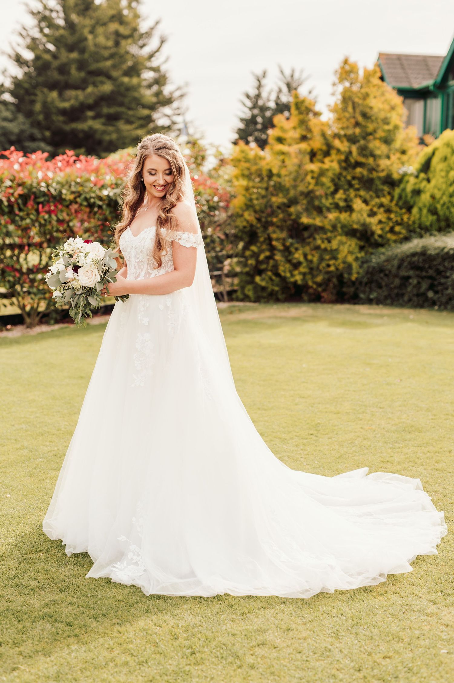 Beautiful wedding dress with flowing tulle train and white floral bouquet photographed outdoors on manicured lawn.