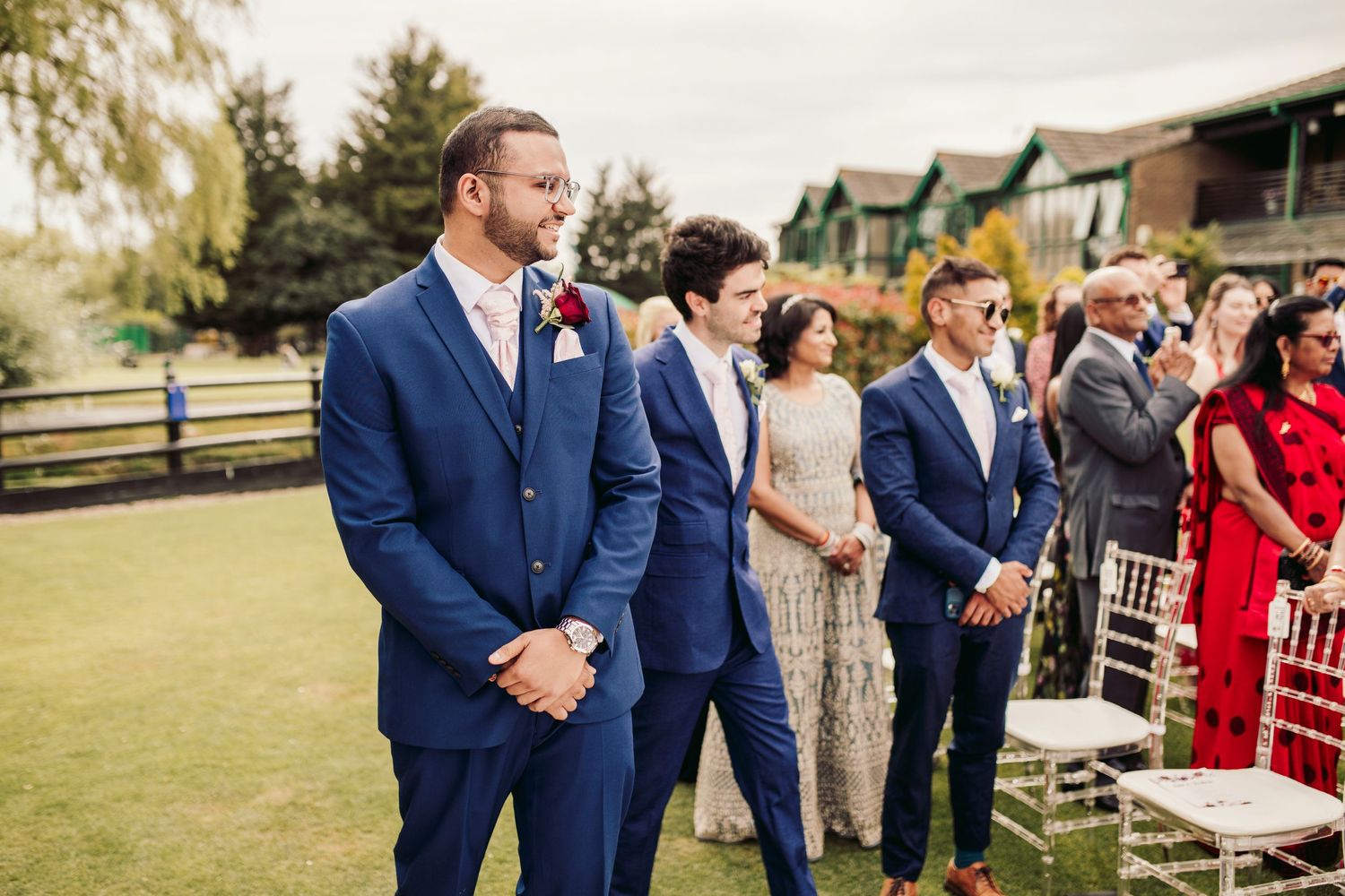 Wedding party wearing navy blue suits and formal attire stands in a row on a grassy lawn at an outdoor venue.