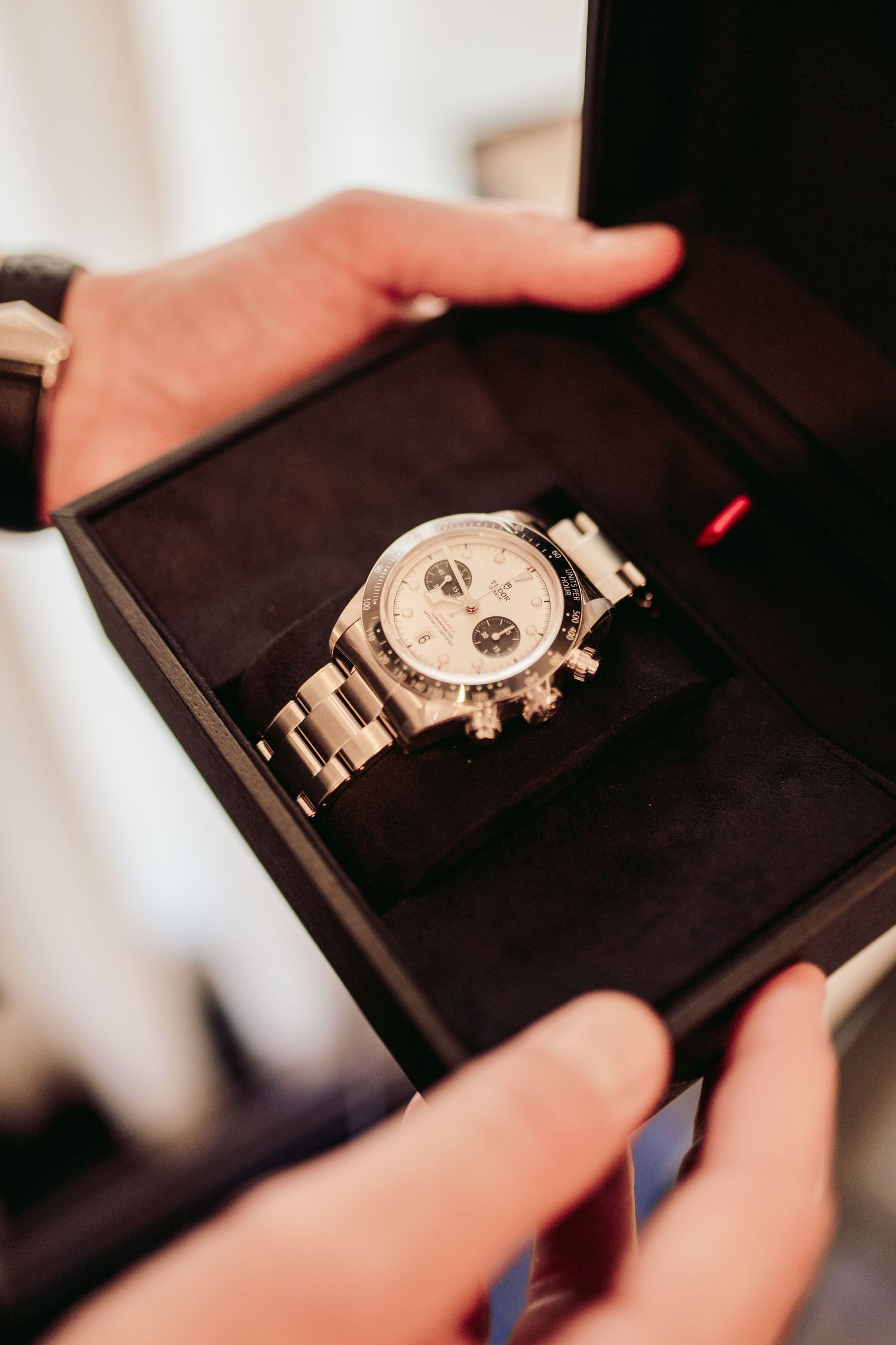 Hands holding open a black jewelry box displaying a silver chronograph watch with metallic bracelet.Claridge’s wedding