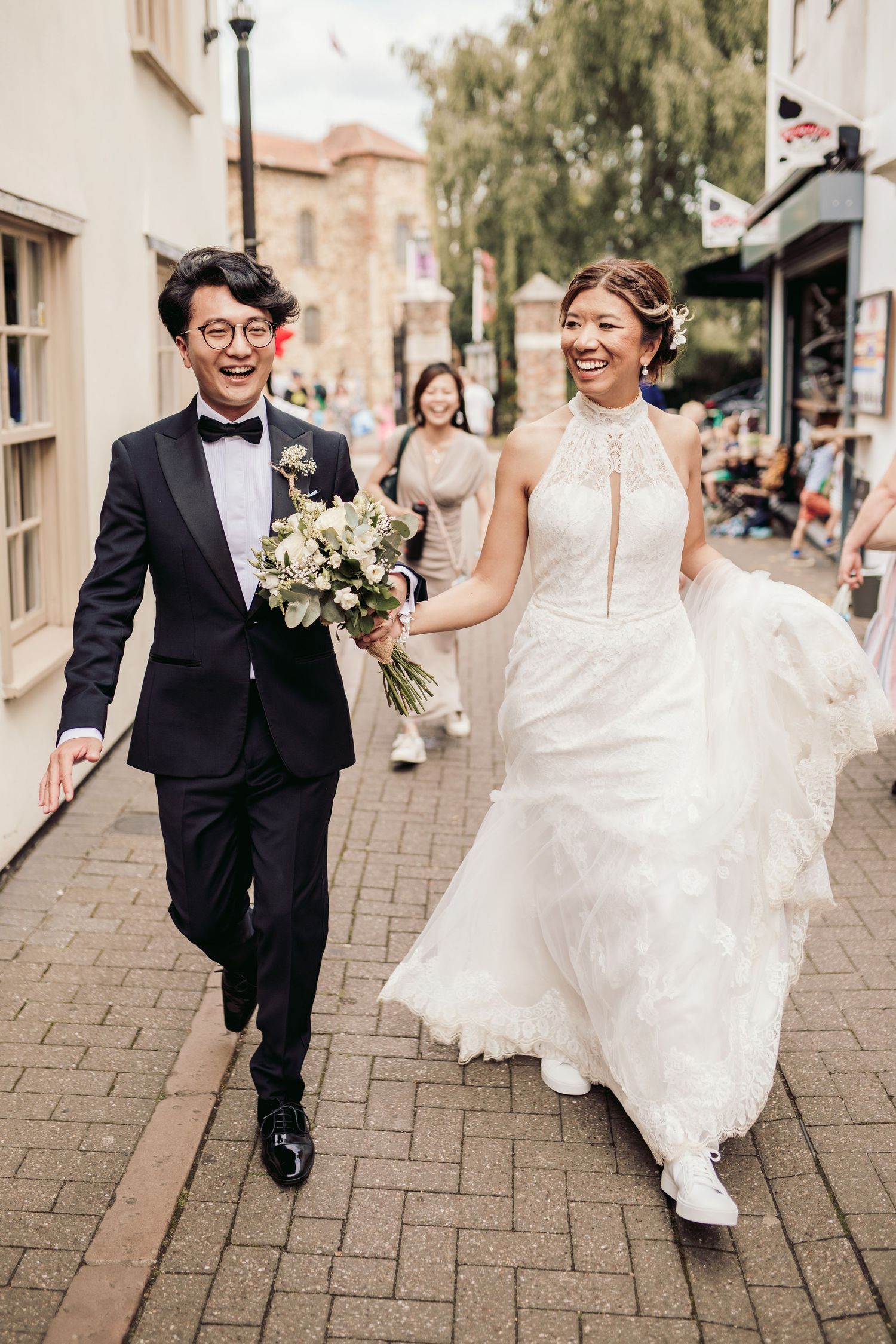 A couple runs playfully along a brick-paved street in formal wedding attire.