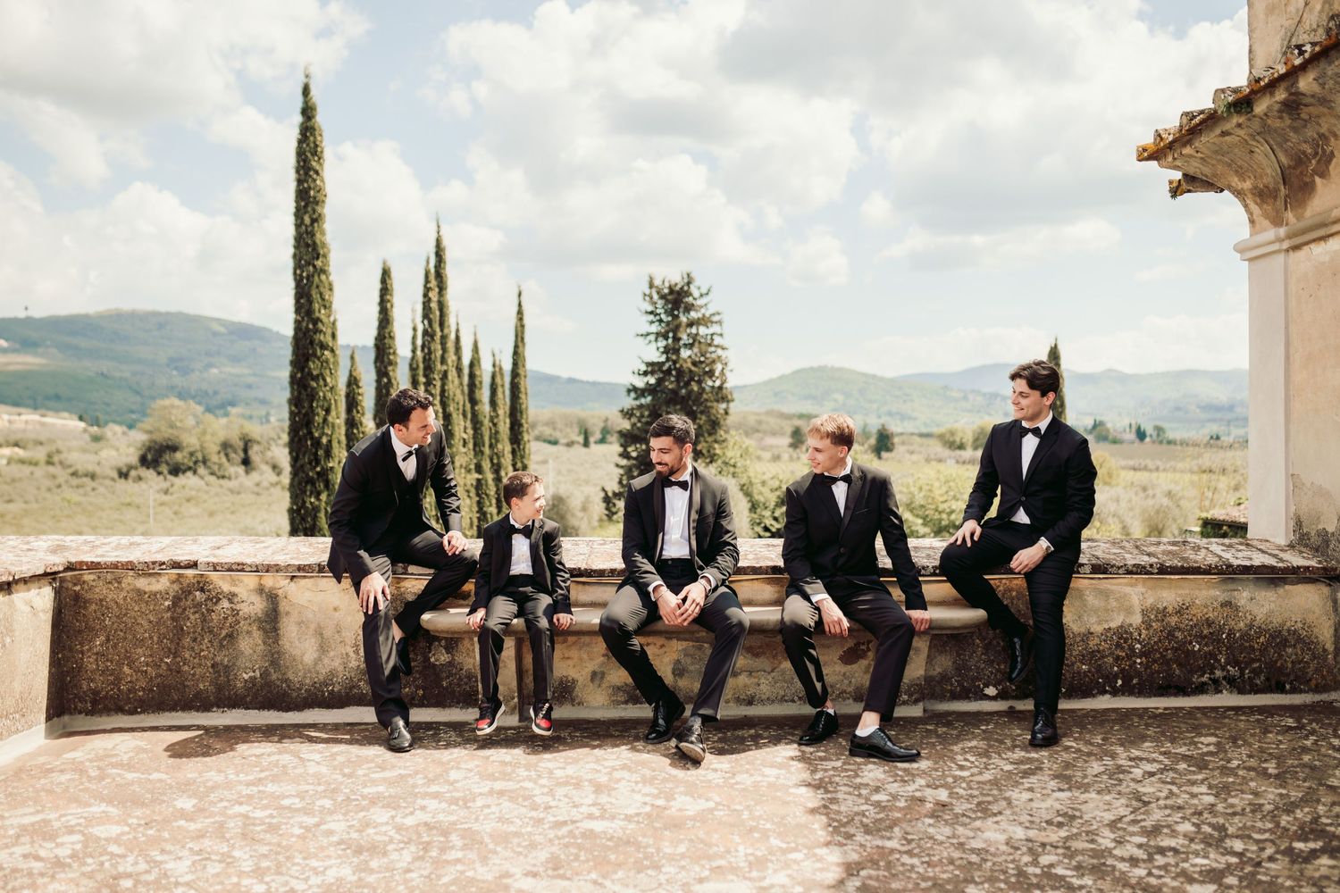 Wedding party members in formal attire lounging on stone wall in Tuscan countryside.