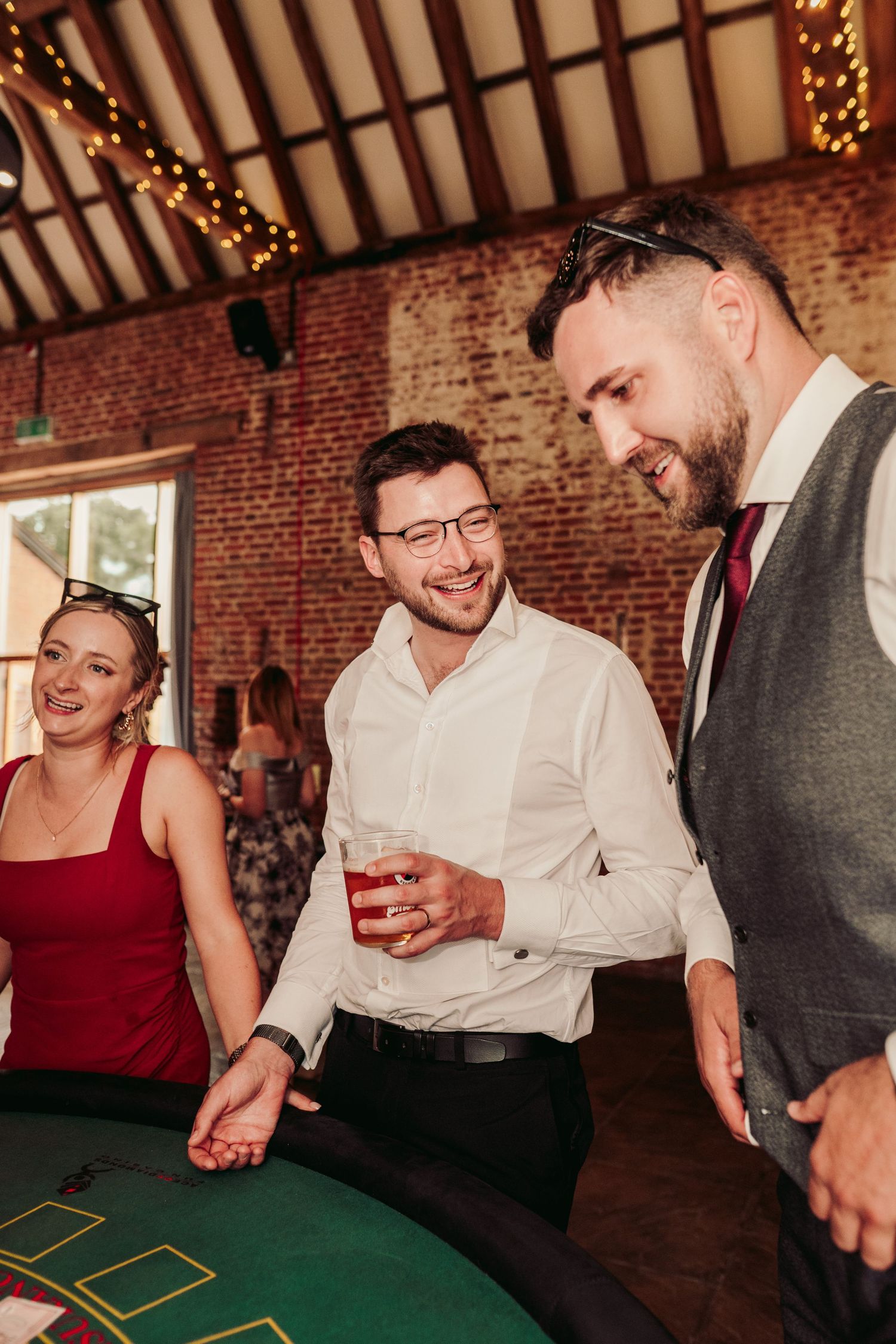 Wedding guests gather around a casino-style gaming table at a reception with exposed brick walls and string lights.