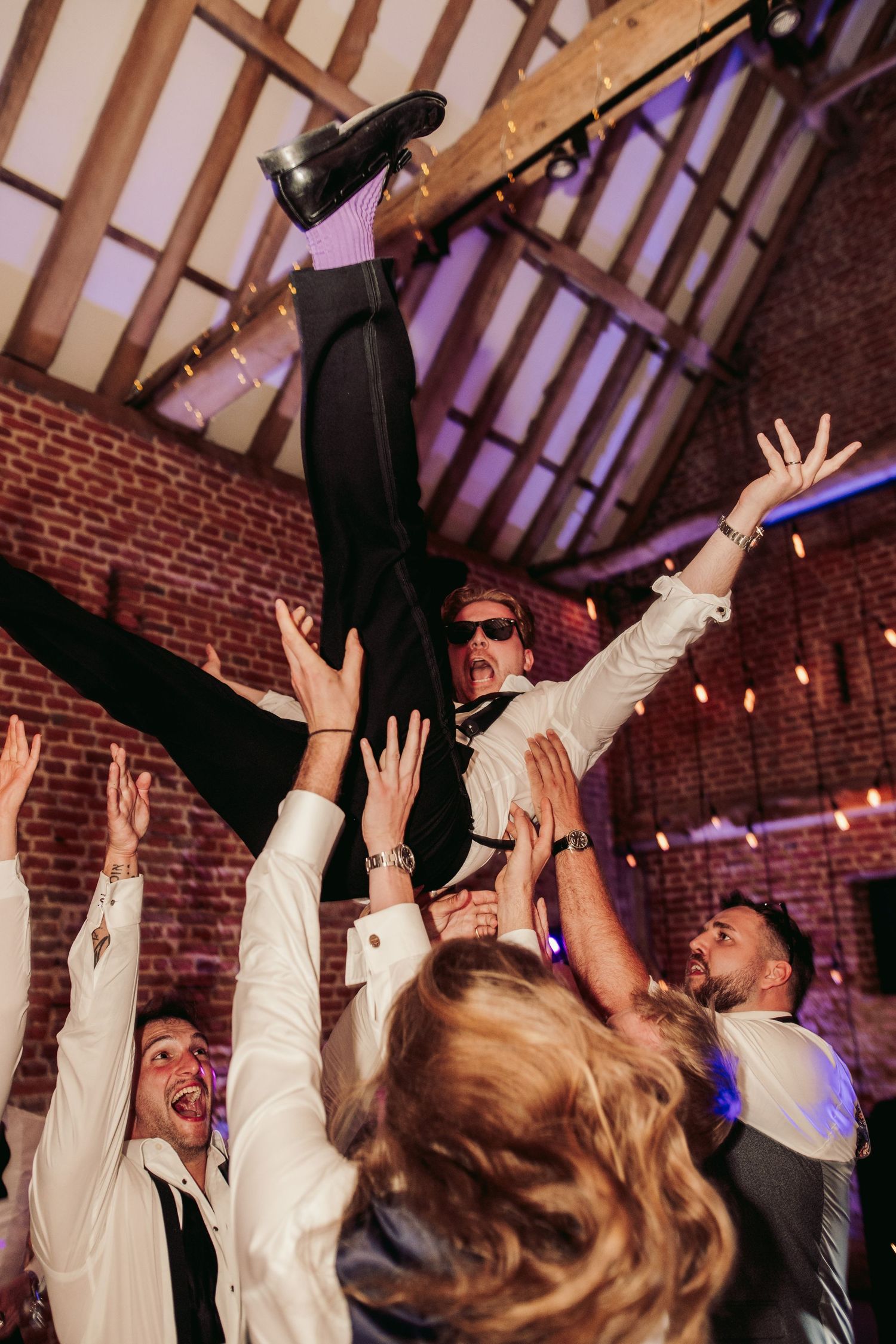 Wedding guests raise their hands in celebration on a dance floor beneath wooden beams and brick walls.