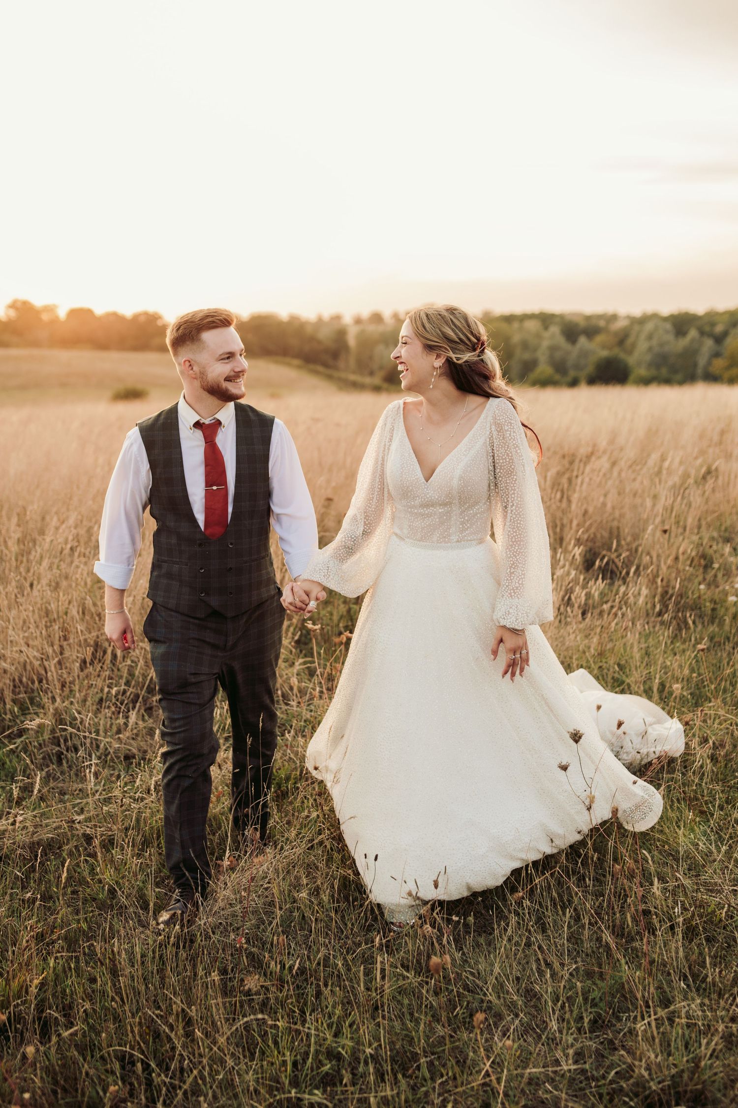 A couple walks hand in hand through a golden wheat field at sunset in wedding attire.