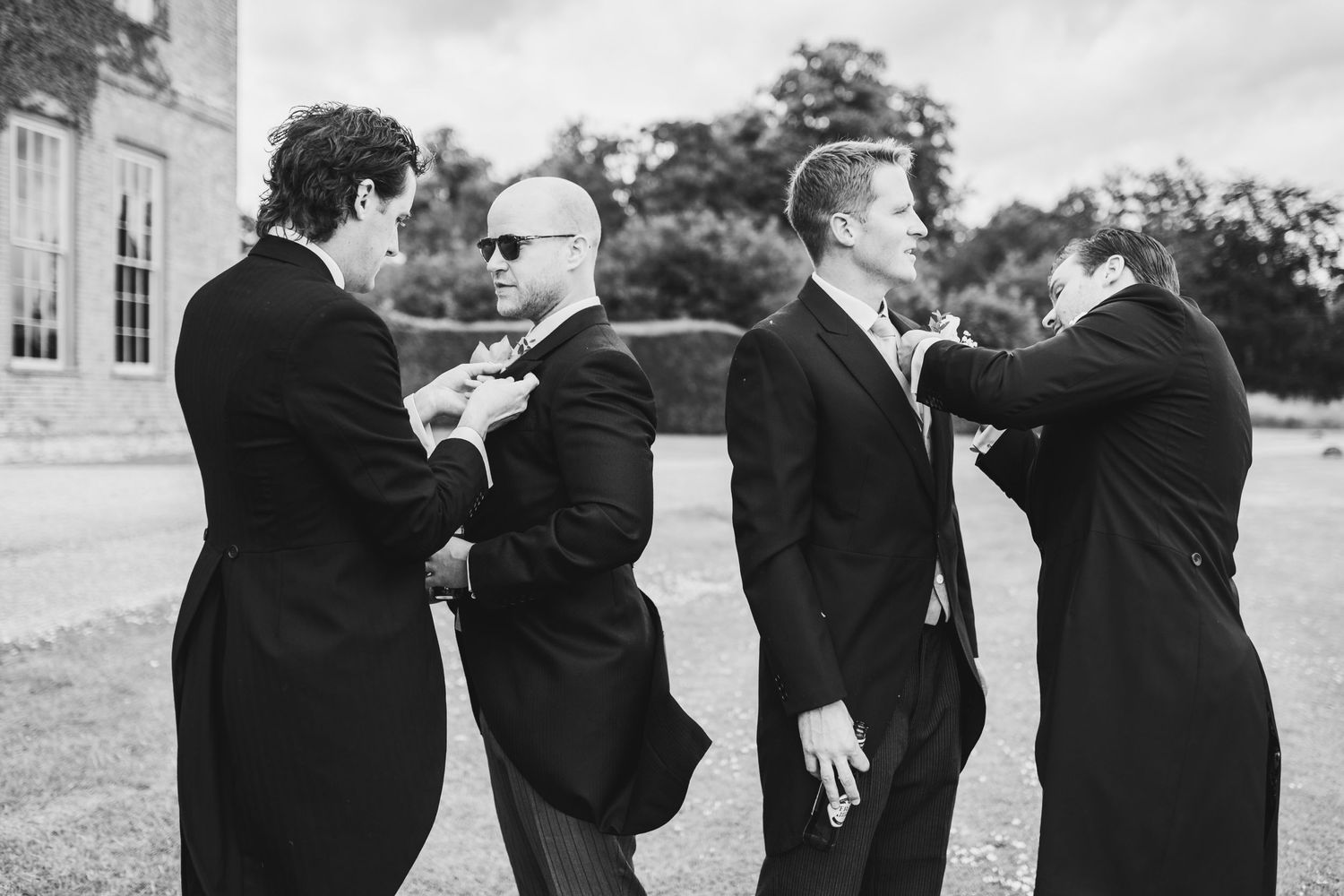 A black and white photo shows four men in formal suits playfully interacting outdoors on a lawn.