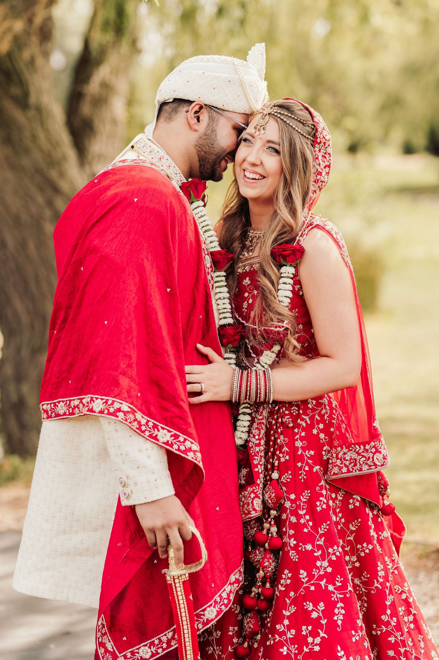 Three Rivers Country Club wedding Couple in traditional red and gold South Asian wedding attire share a joyful moment outdoors under trees.