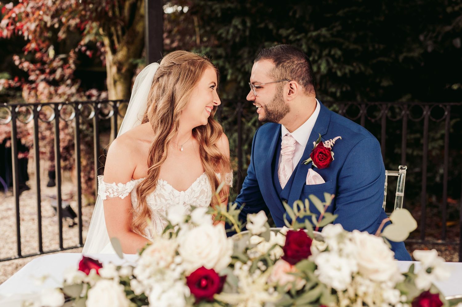 Newlyweds share a joyful moment at their reception table decorated with red and white floral arrangements.