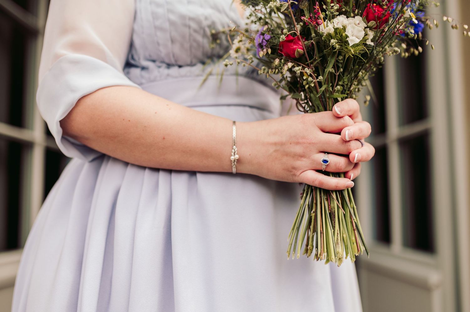 Someone wearing a delicate bracelet holds a rustic bouquet with red and white flowers against a light gray dress. Spiez switzerland