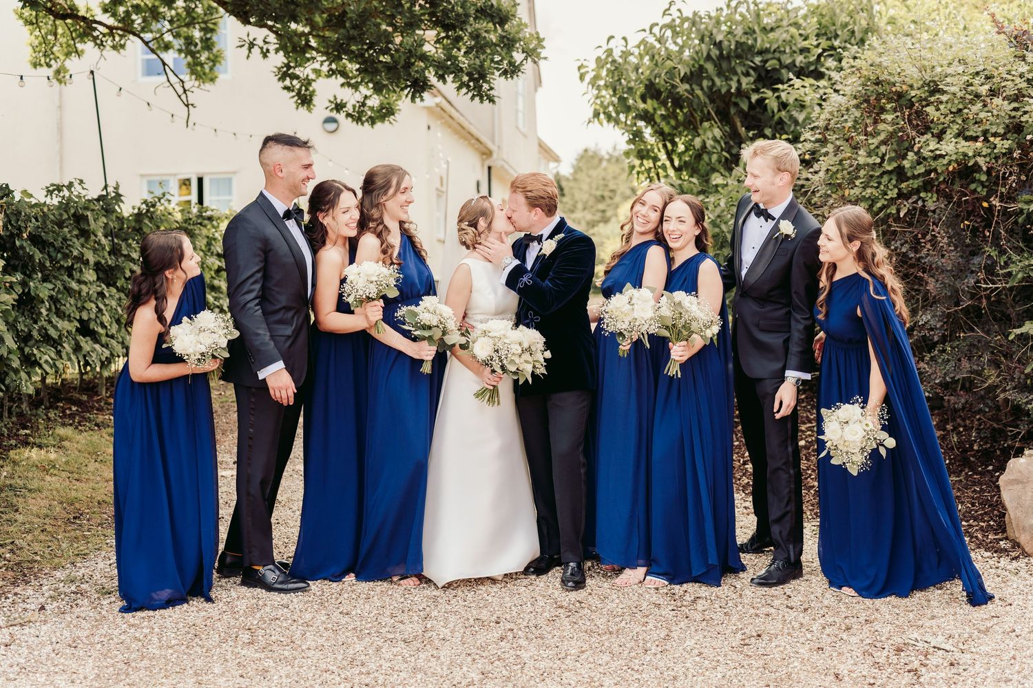 Wedding party in coordinated royal blue and black attire poses together outdoors on a sunny day.