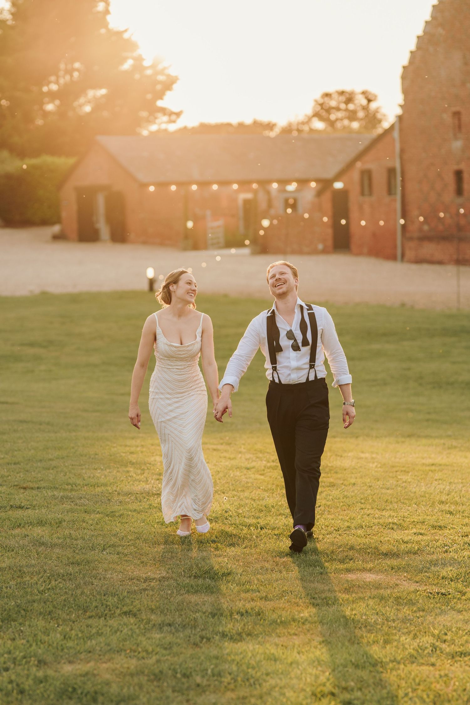A couple walks hand in hand through a sunlit grassy field at golden hour during their wedding celebration.
