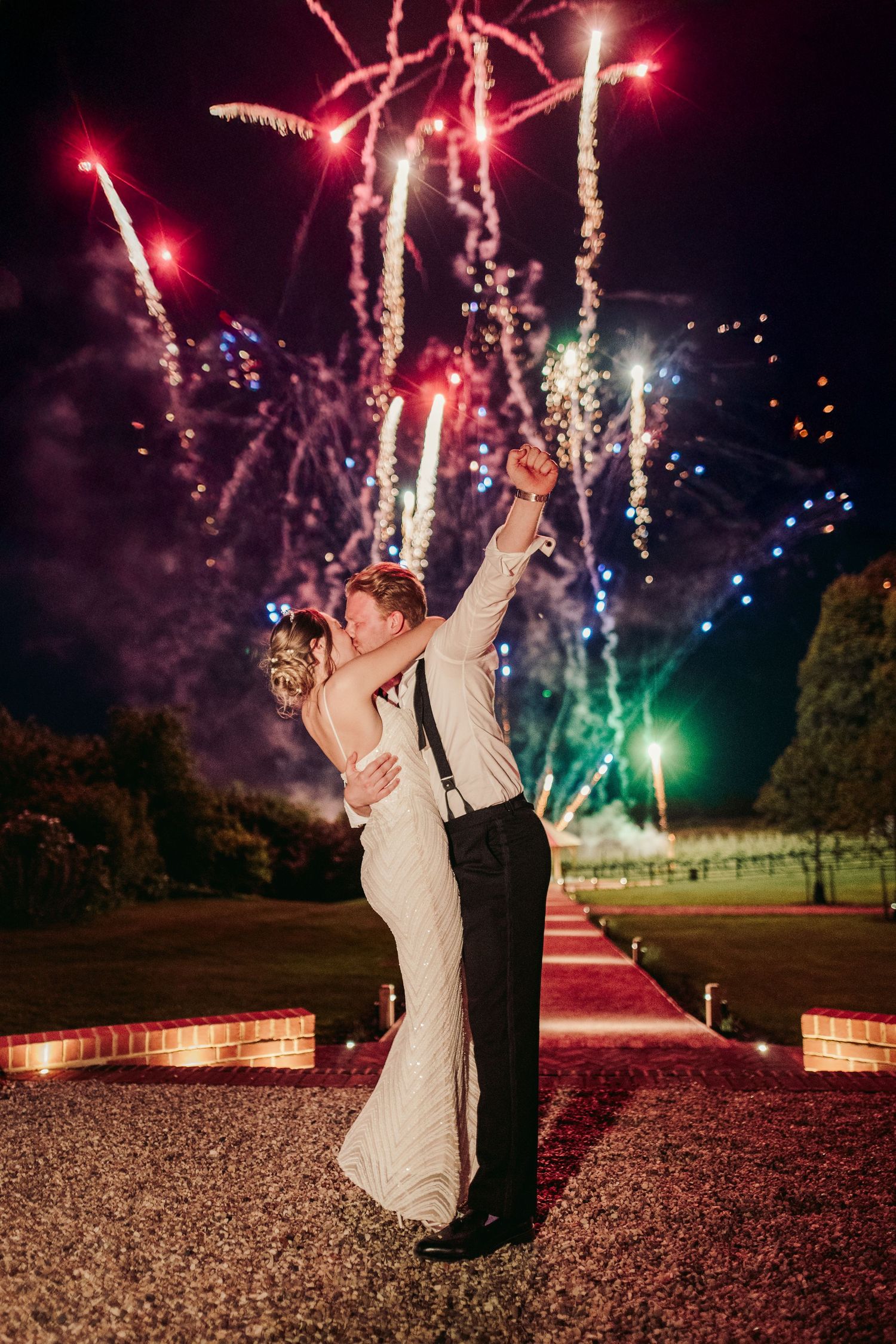 Couple dances under colorful fireworks display at nighttime wedding celebration on brick patio.