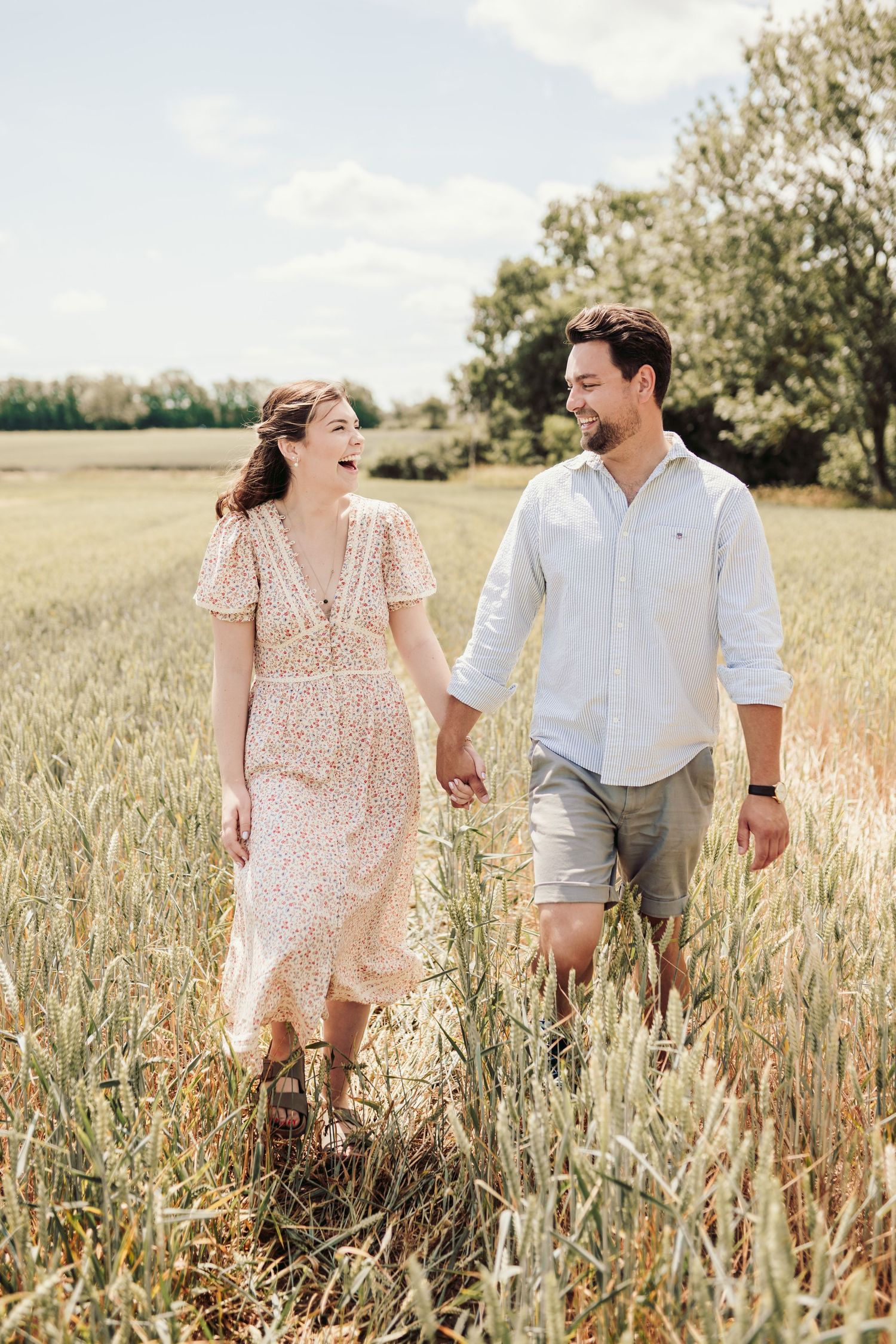 A couple walks hand in hand through a golden wheat field at sunset wearing casual summer attire.