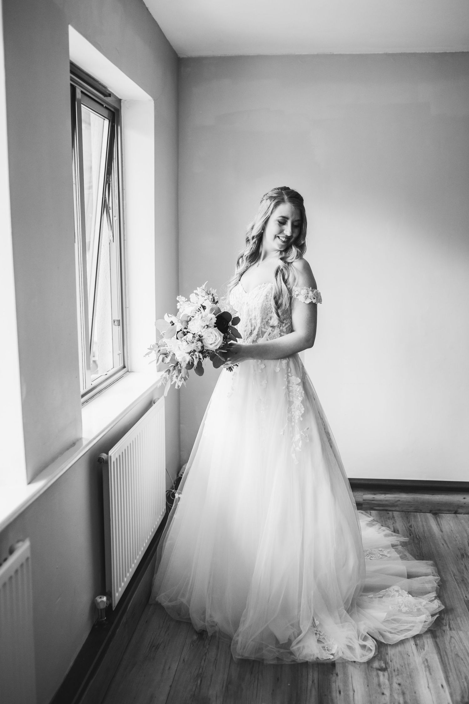 Black and white photo of a bride in a flowing lace gown holding a floral bouquet while standing by a window.