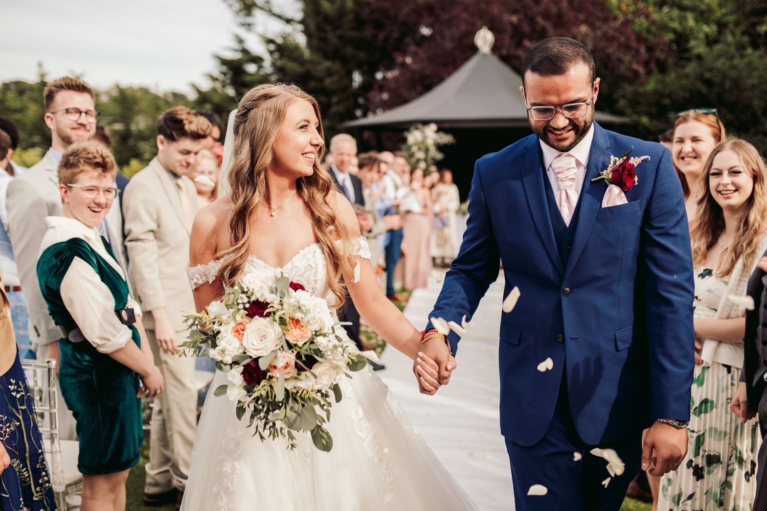 A joyful couple walks hand in hand during outdoor wedding ceremony while guests smile in the background.
