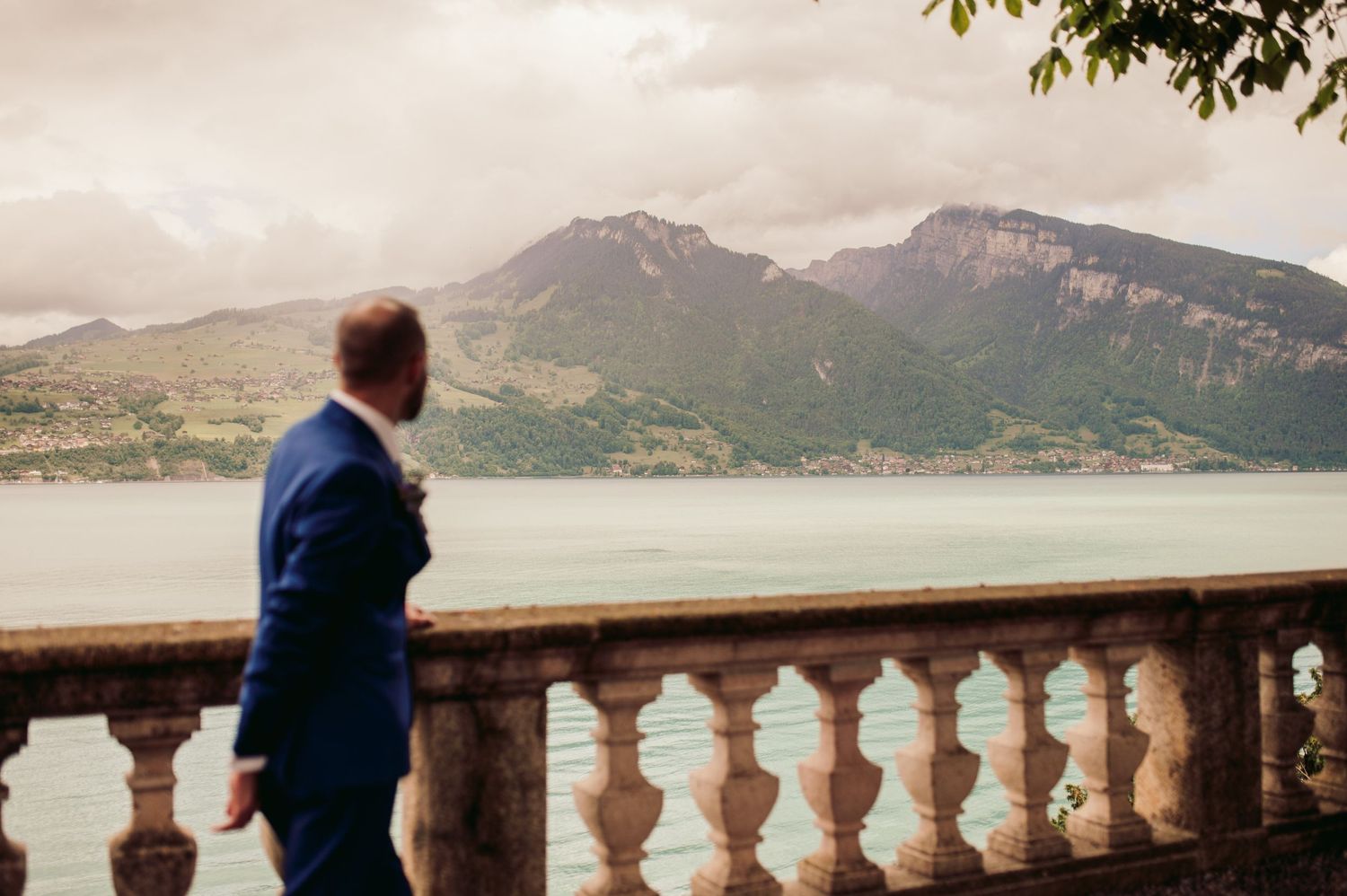 Someone in a blue suit admires the scenic mountain lake view from an ornate stone balustrade overlook. Spiez switzerland destination wedding