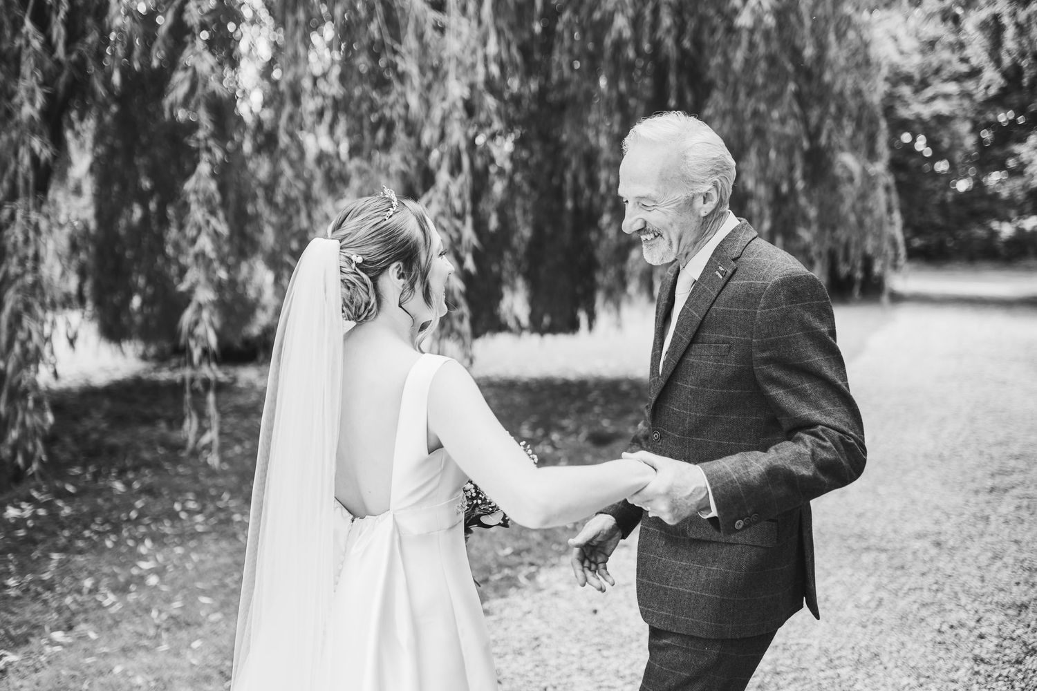 A couple shares a romantic moment in a garden setting with trees and a long white veil flowing in the breeze.