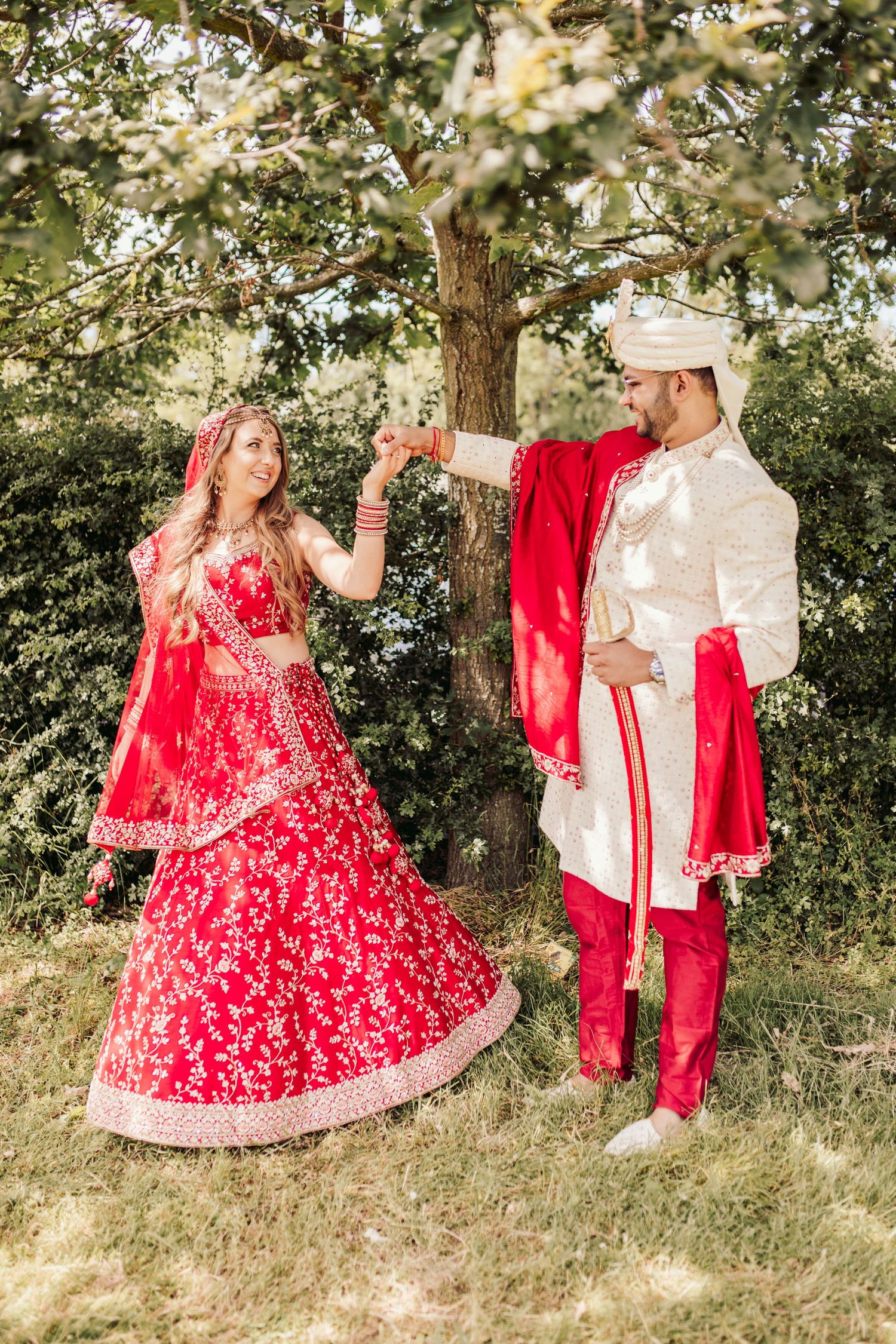 A couple in traditional South Asian wedding attire poses outdoors under a tree in red and white formal clothing.