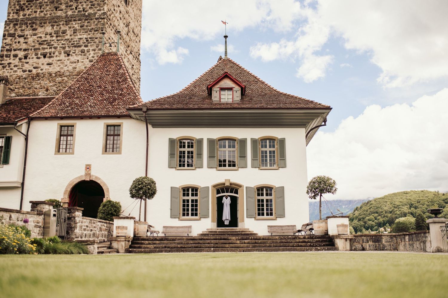 White historic manor house with brown roof, green lawn and stone tower stands elegantly against cloudy sky. Spiez switzerland destination we