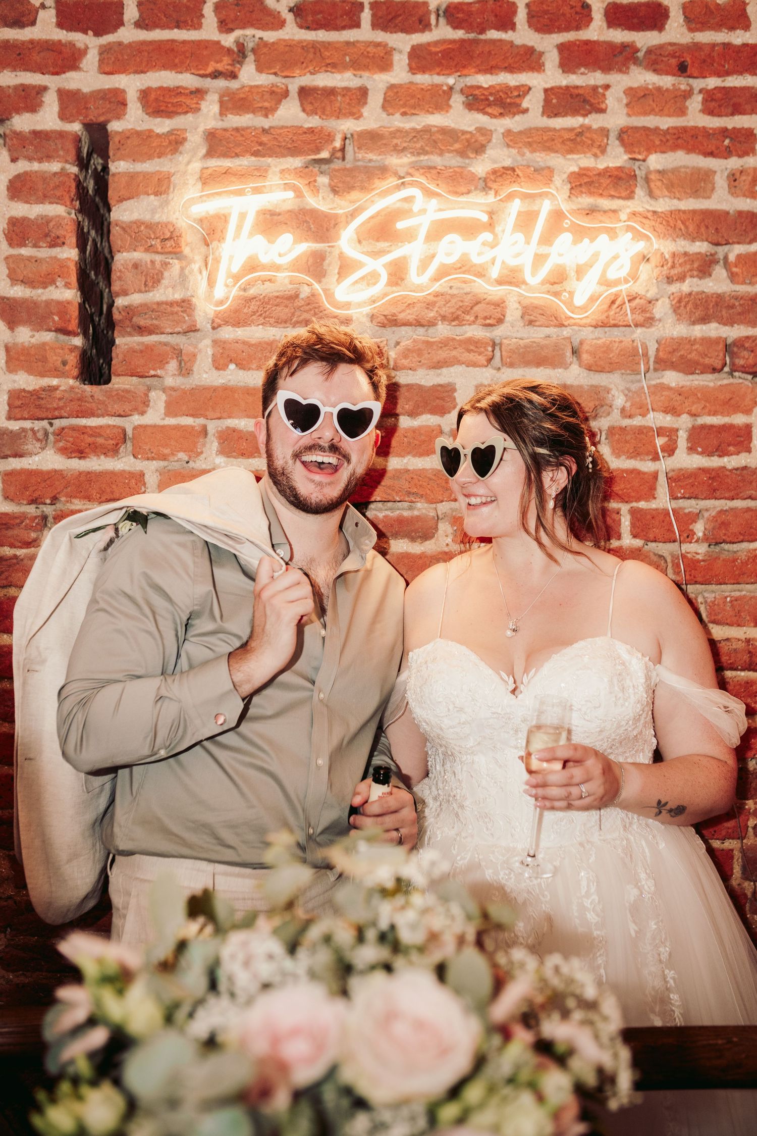 Wedding couple wearing heart-shaped sunglasses against brick wall with neon sign.