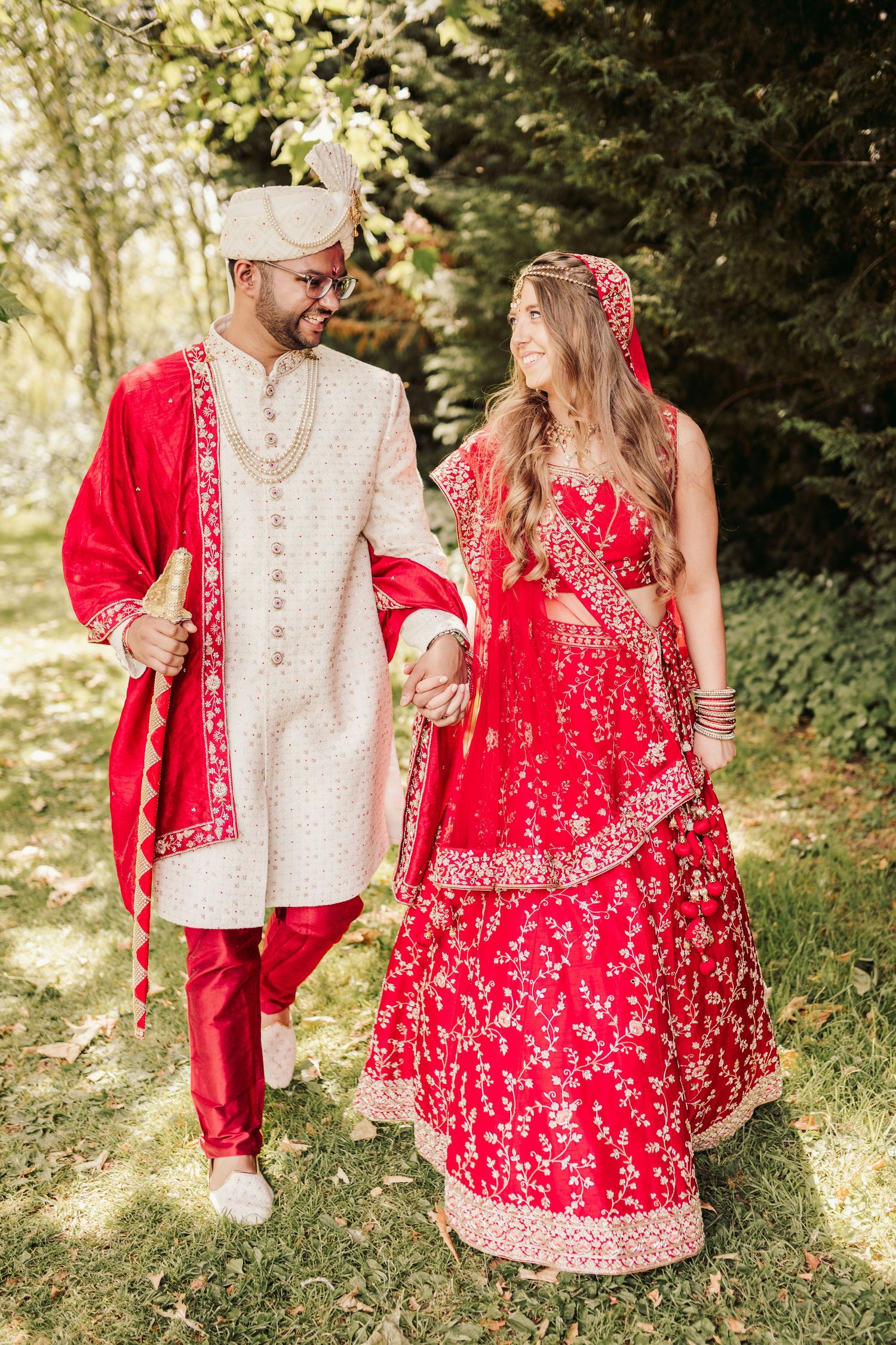 A couple wearing traditional Indian wedding attire - red and gold lehenga and cream sherwani with red dupatta walking outdoors.