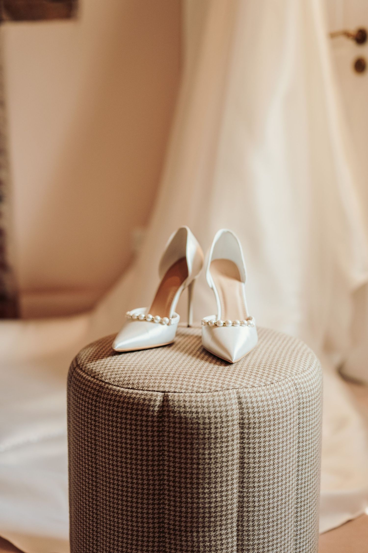 Elegant white bridal heels with pearl details sit atop a gray textured ottoman against a light backdrop.