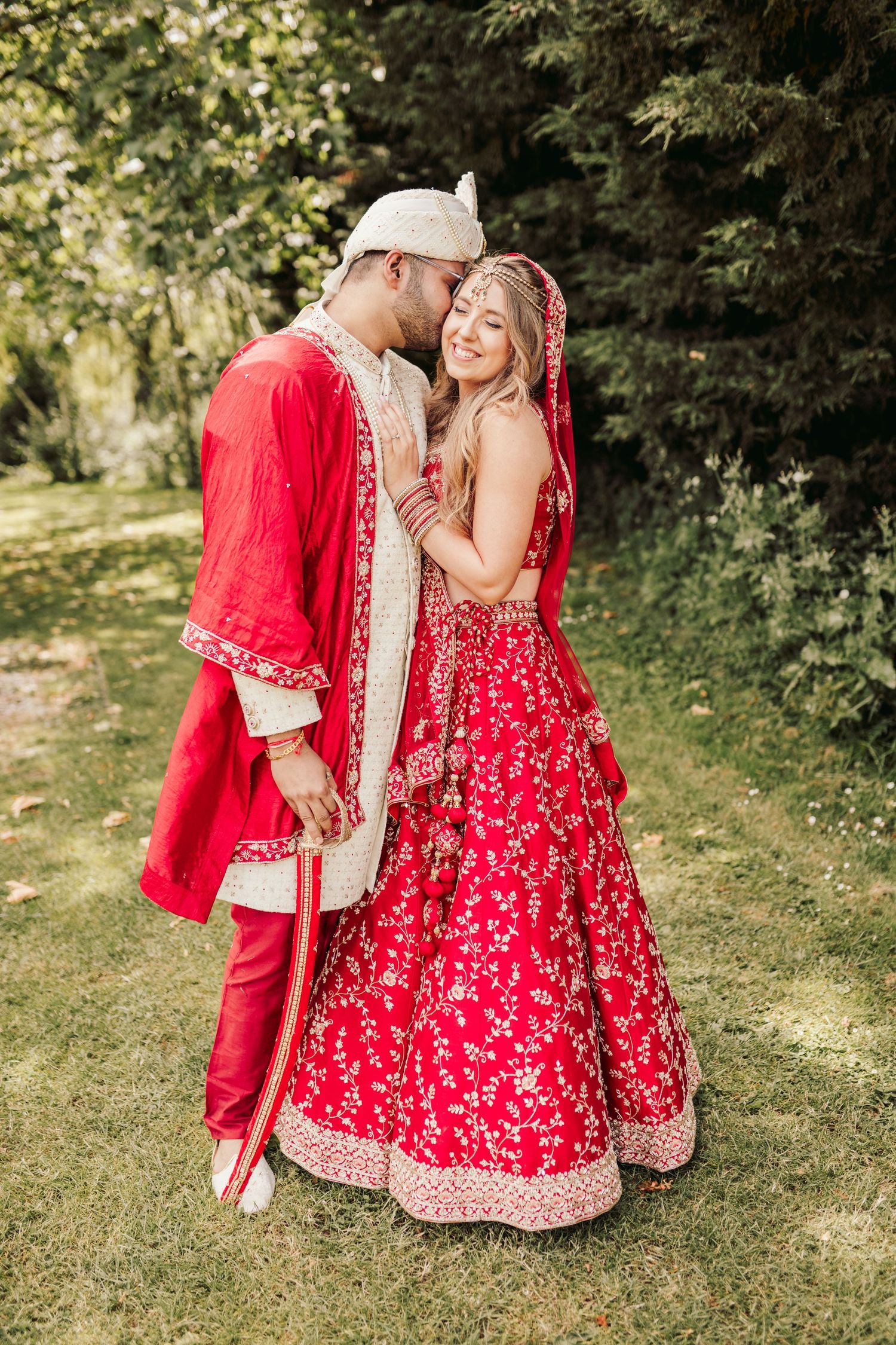 A couple in traditional South Asian wedding attire embraces outdoors, wearing red and cream ceremonial garments.