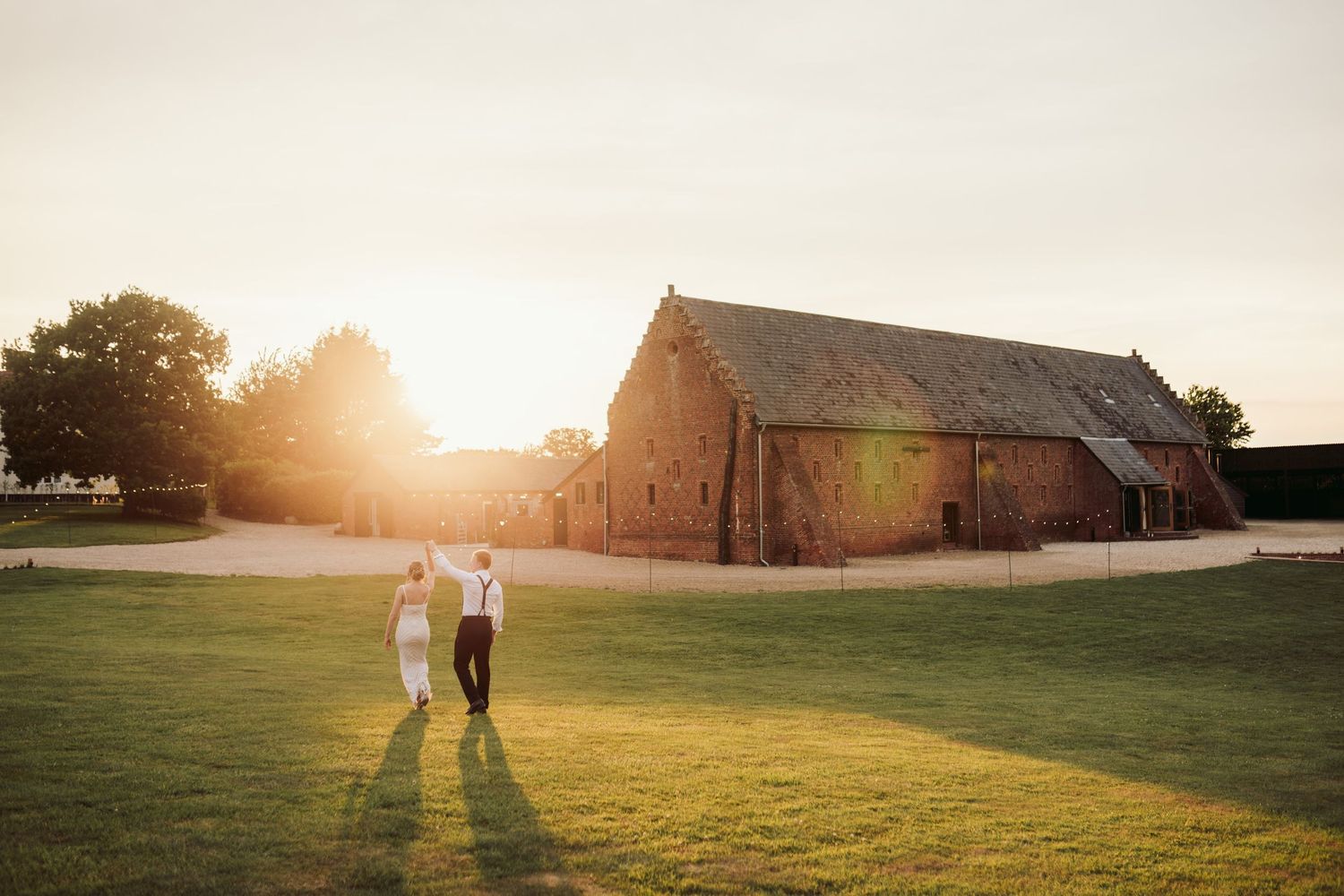Couple walking hand in hand through a sunlit meadow at sunset with rustic barns in the background.