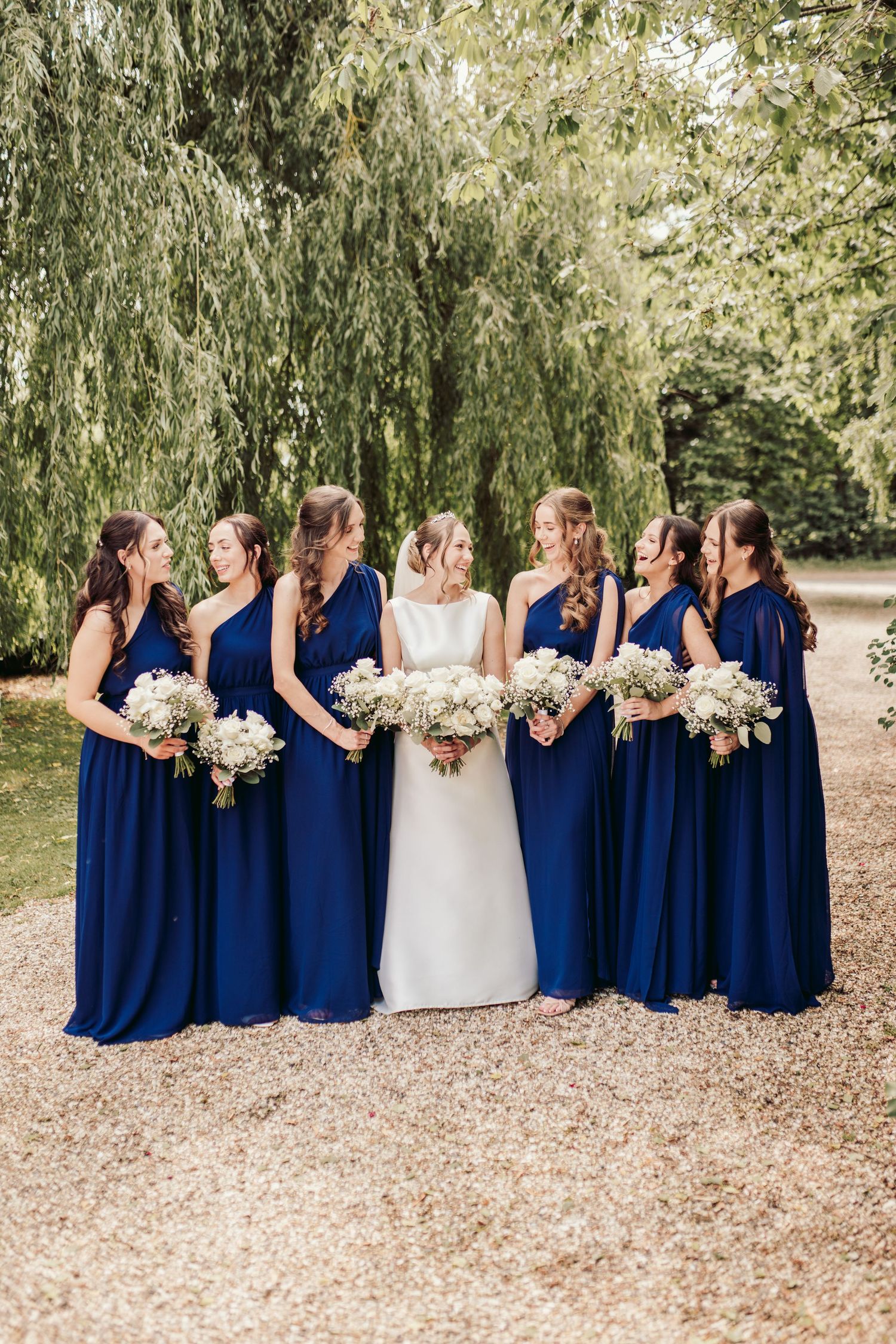 Wedding party in navy blue bridesmaid dresses and white bridal gown pose in line holding floral bouquets outdoors.
