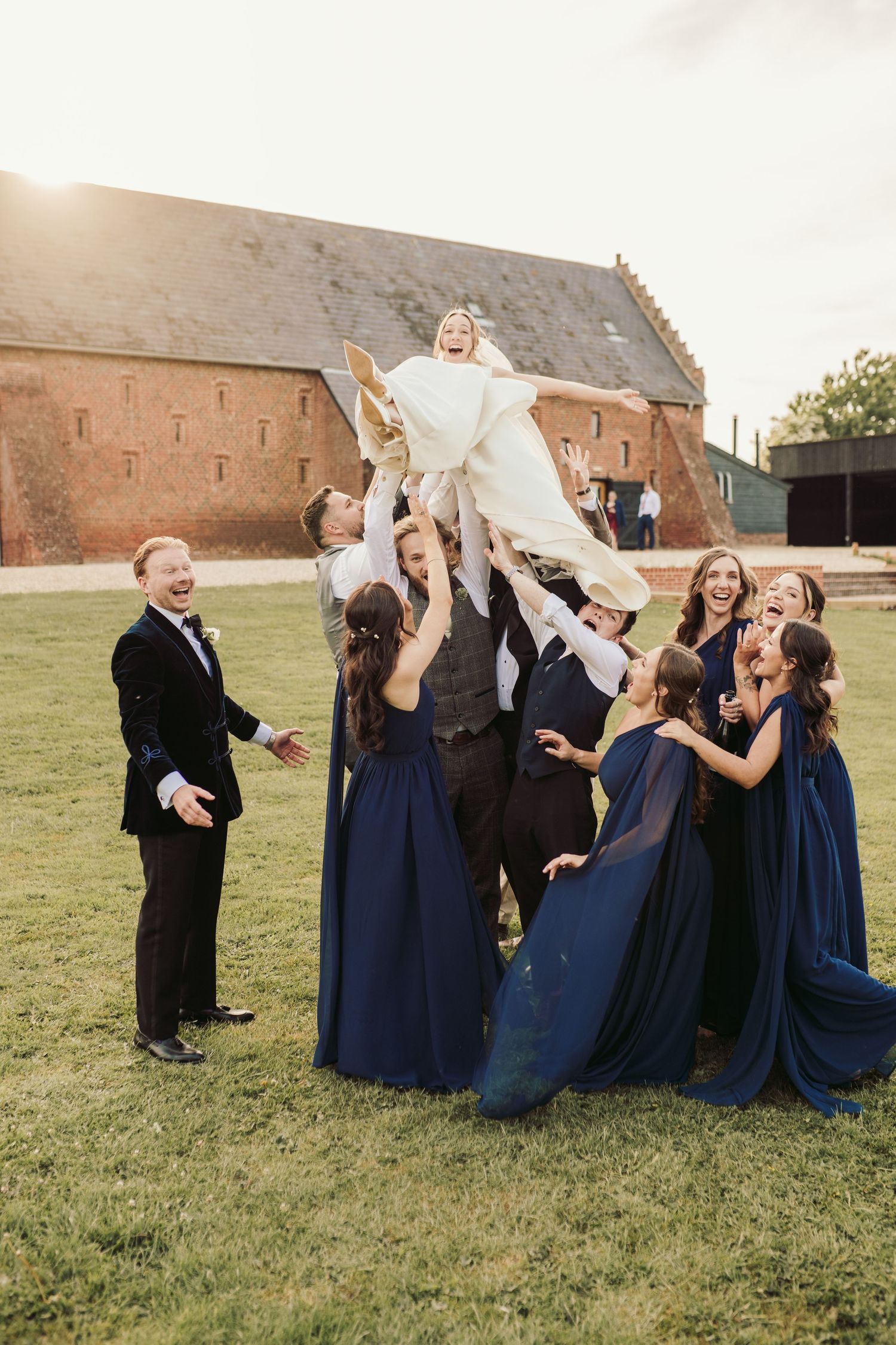 Wedding party in navy blue dresses playfully toss someone in a white dress into the air on a lawn near a brick building.