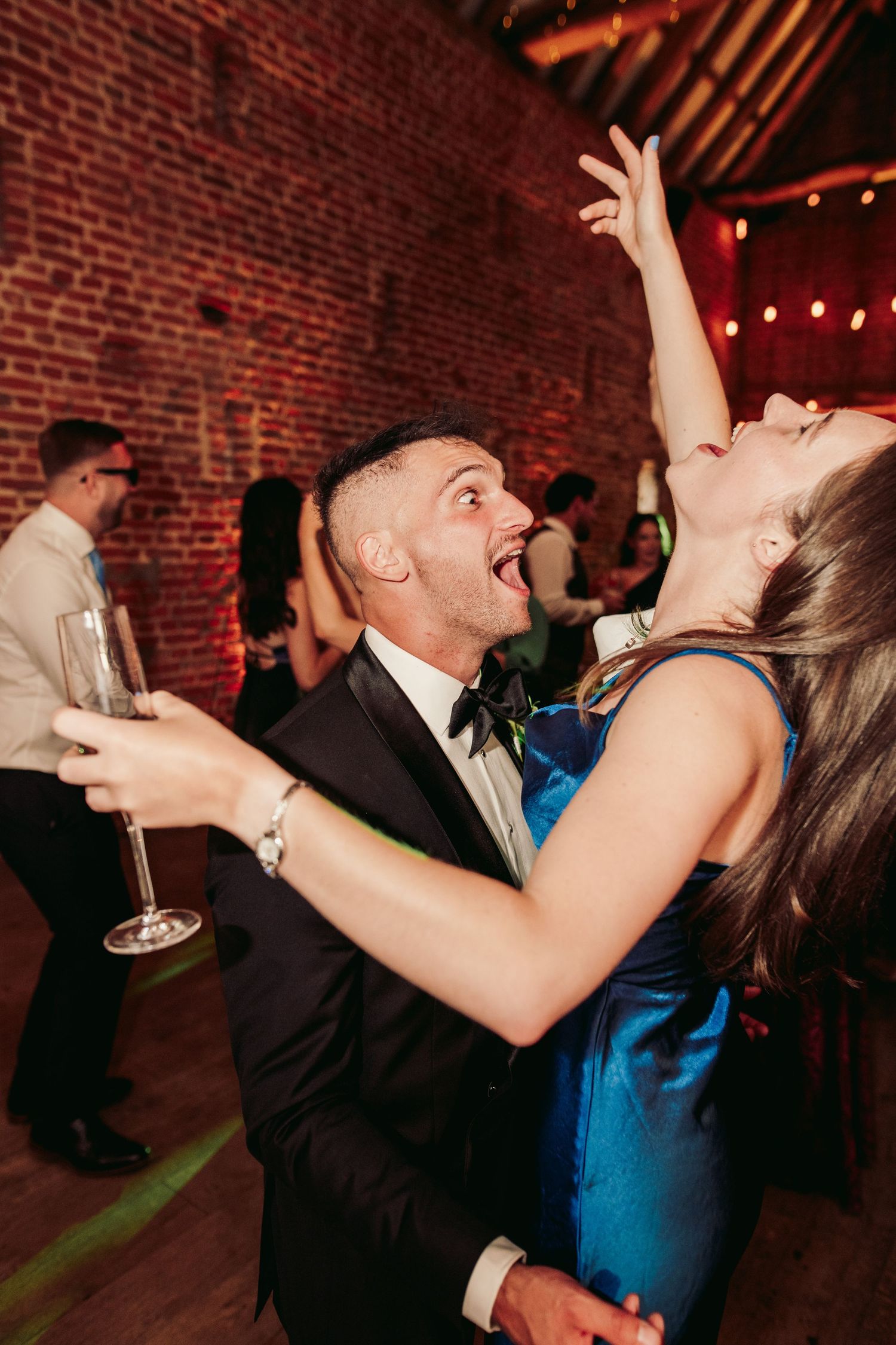 Couples dancing and celebrating at a rustic brick-walled venue with string lights overhead during a festive party.