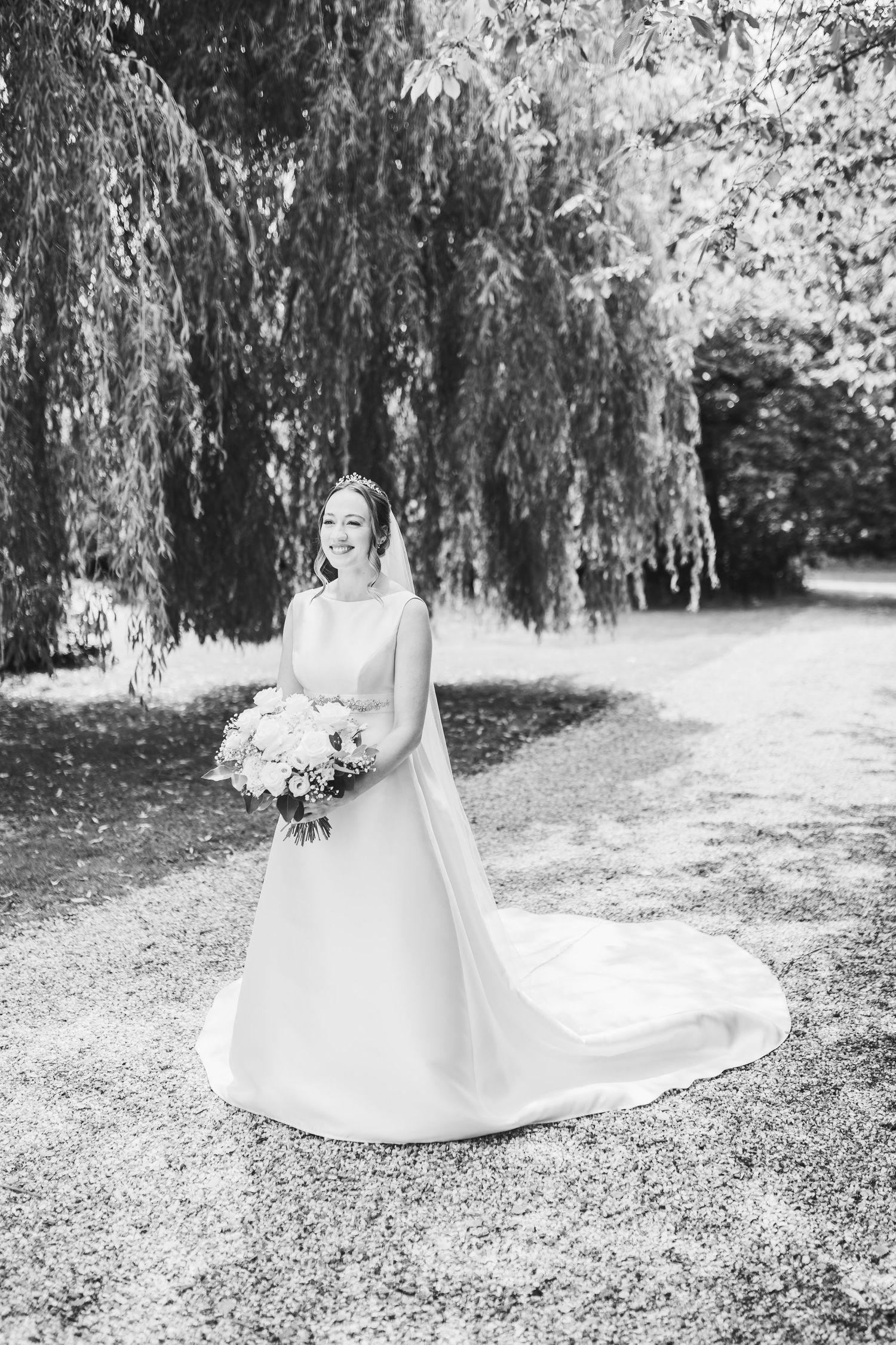 A bride in a flowing white wedding dress sits gracefully under a weeping willow tree in a black and white photograph.