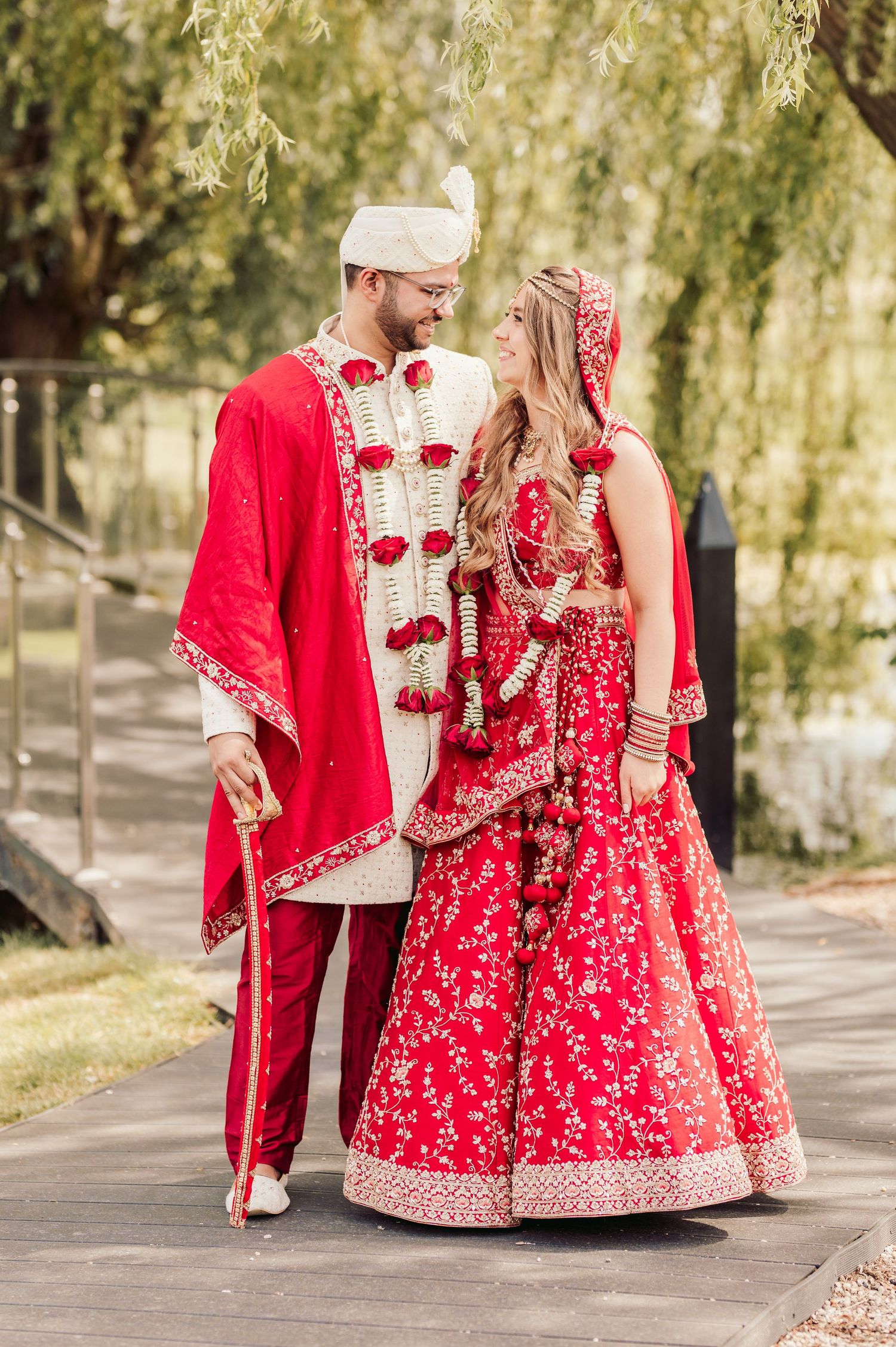 A couple in traditional South Asian wedding attire stands together on a pathway, wearing matching red and white embellished outfits.