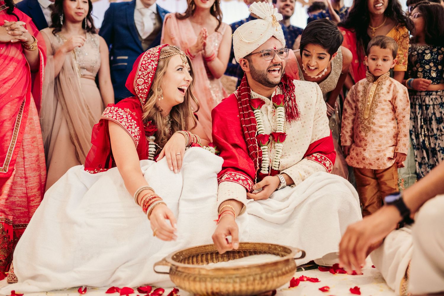 A couple in traditional red and white wedding attire participates in a cultural ceremony surrounded by family and guests.